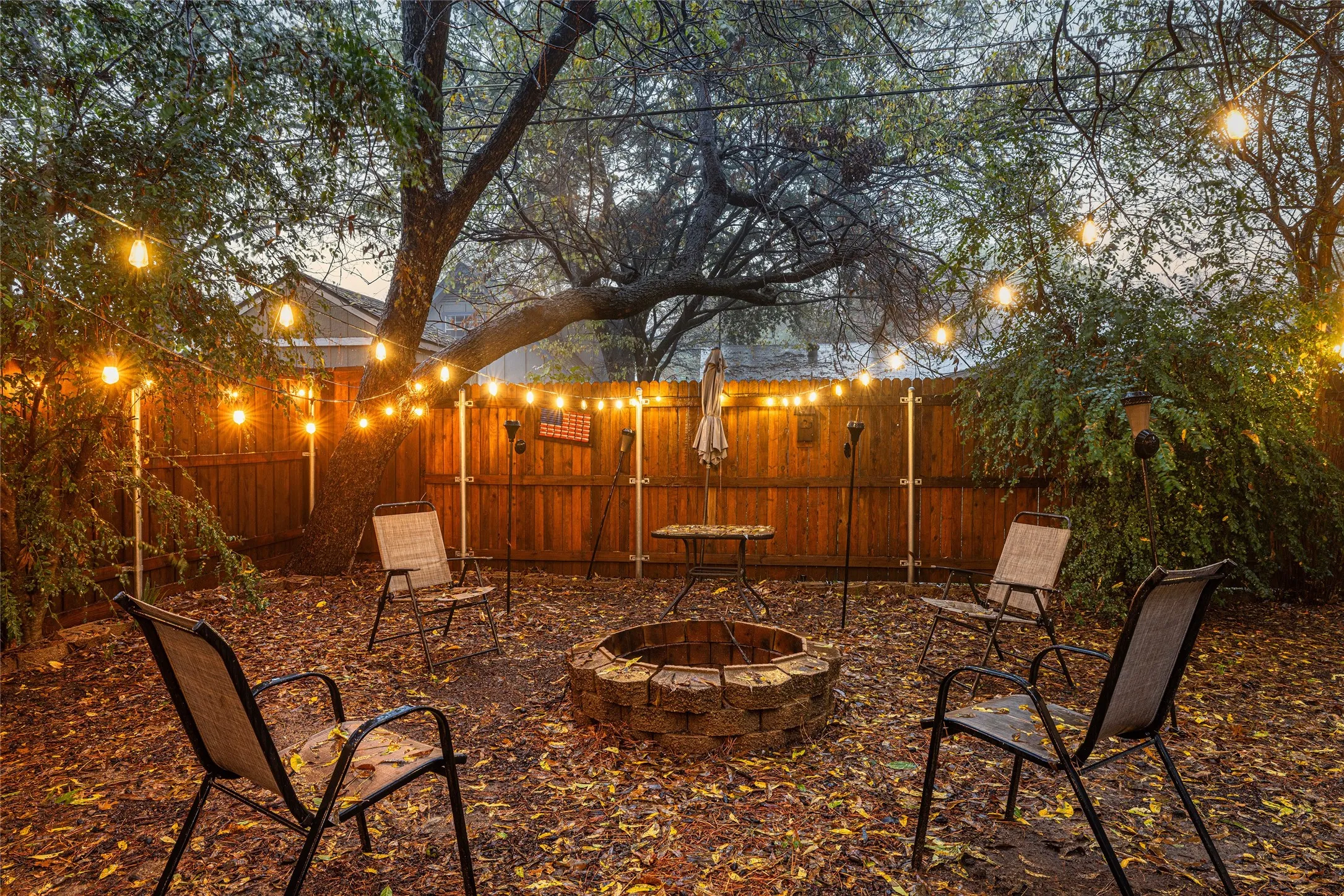 Fenced backyard with a built-in stone fire pit, mature trees, and string lighting