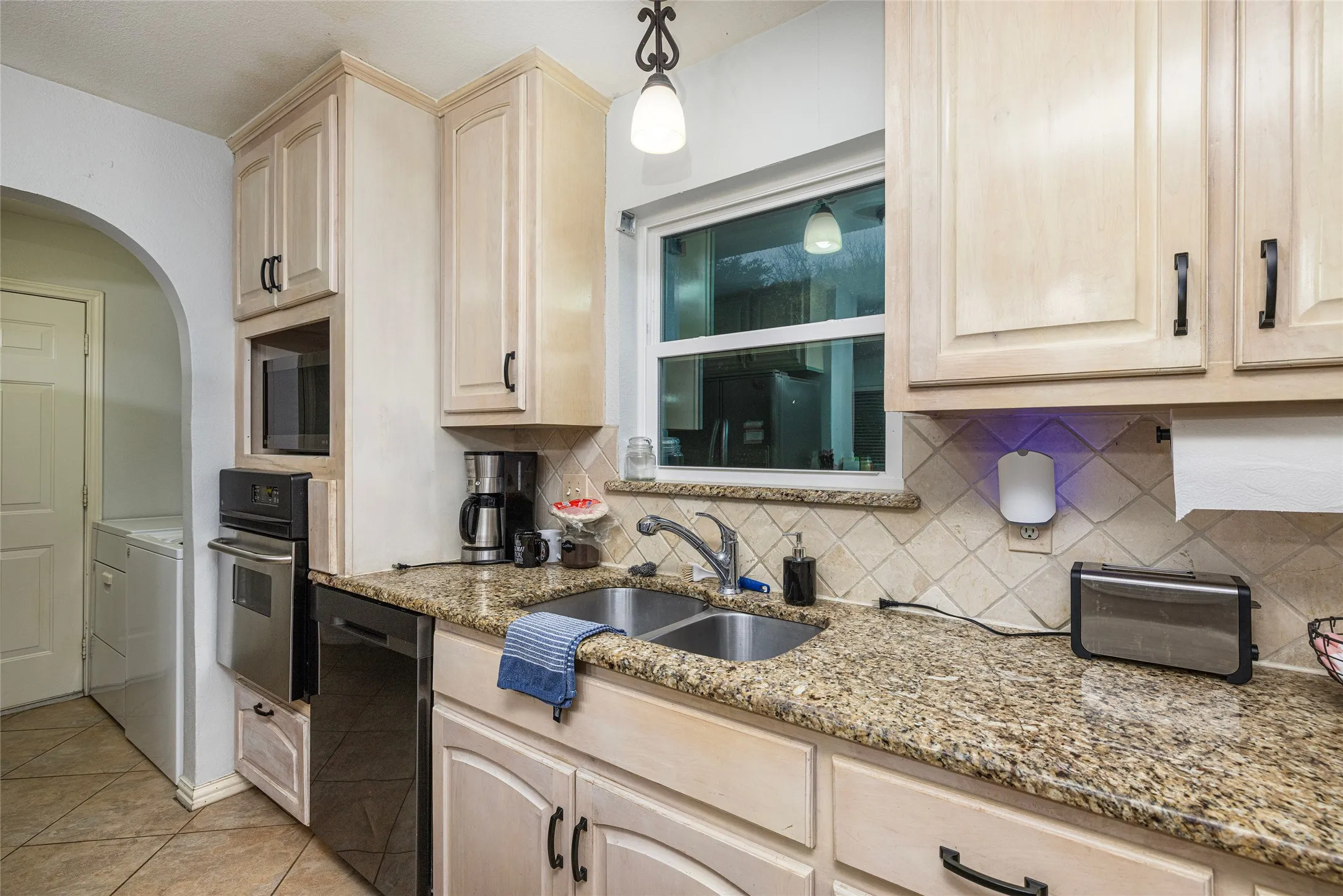 Light wood-finish cabinetry with black hardware, granite-style countertops, stainless steel appliances, and a diagonal tile backsplash