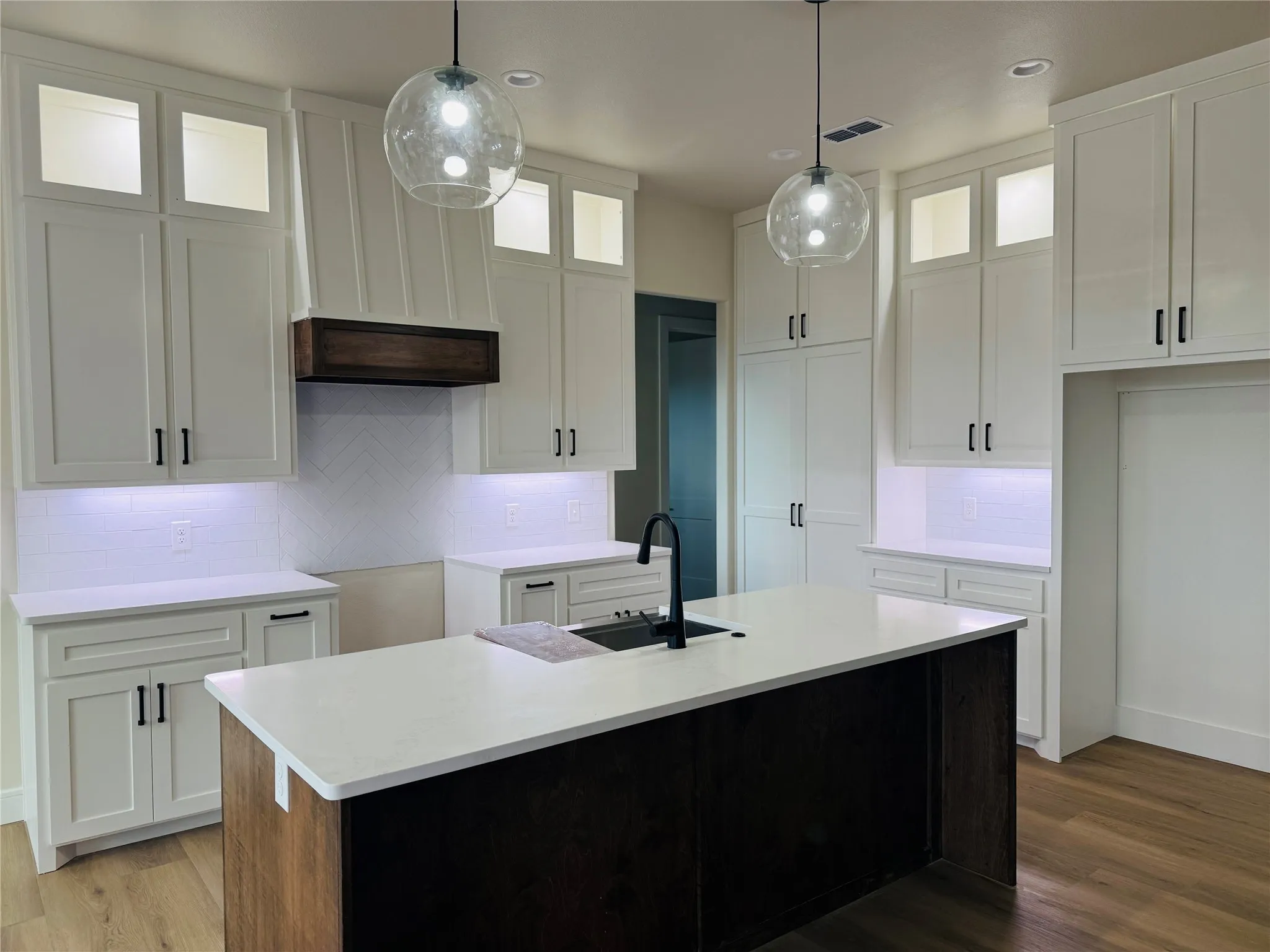 Contemporary kitchen featuring white shaker-style cabinetry with brushed nickel hardware, white quartz countertops, a black waterfall-edge island with wood-finish base, pendant lighting with clear glass globes, and under-cabinet LED accent lighting