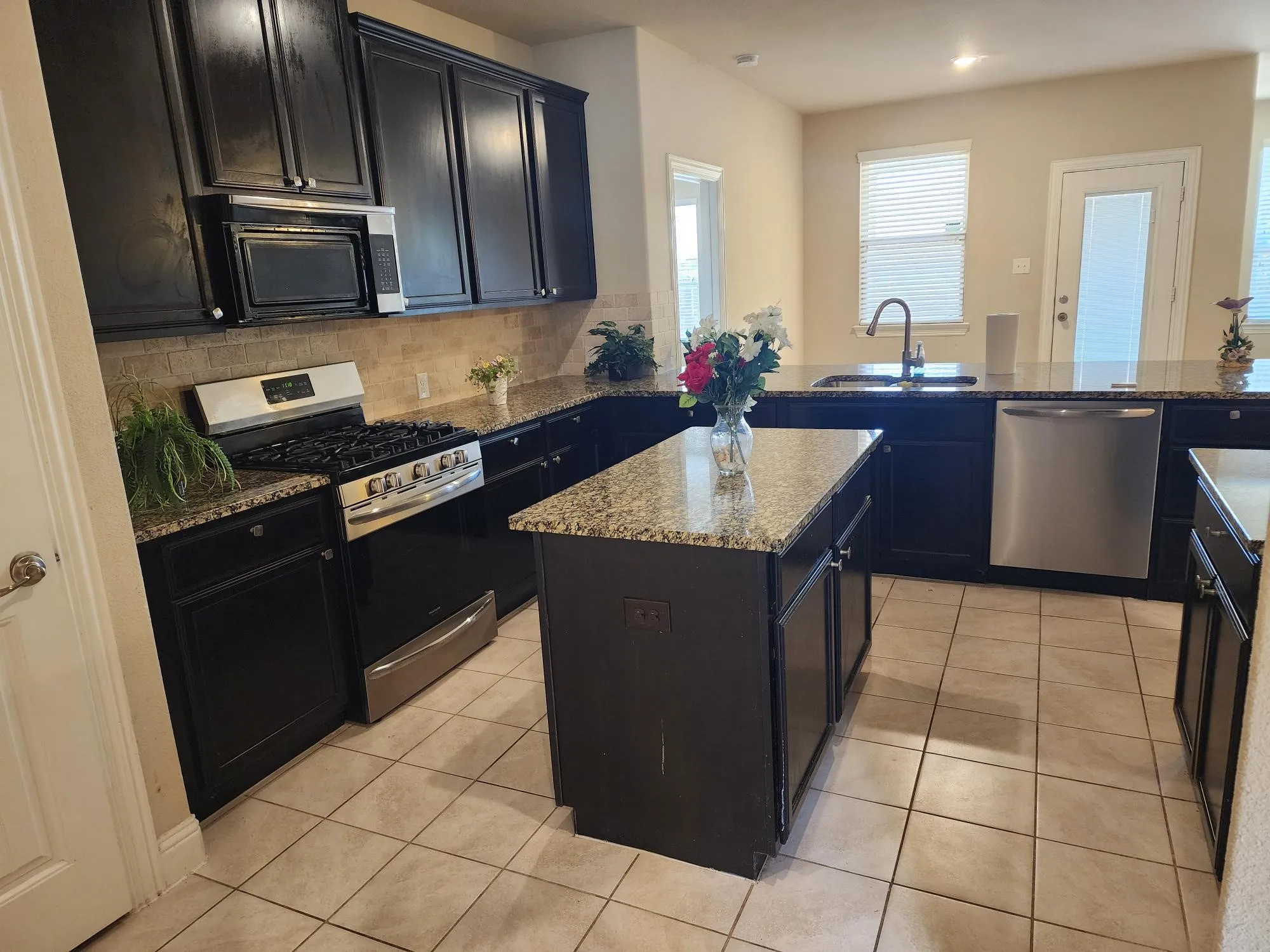 Chef's kitchen featuring dark cabinetry, stainless steel appliances including a gas range and dishwasher, granite countertops, a large center island with seating, tile flooring, and abundant natural light from multiple windows and glass doors