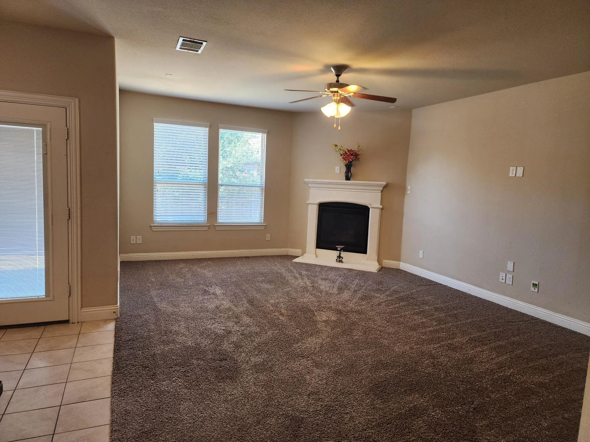 Spacious living area featuring plush carpeting, a built-in fireplace with mantel, ceiling fan, and multiple windows with blinds