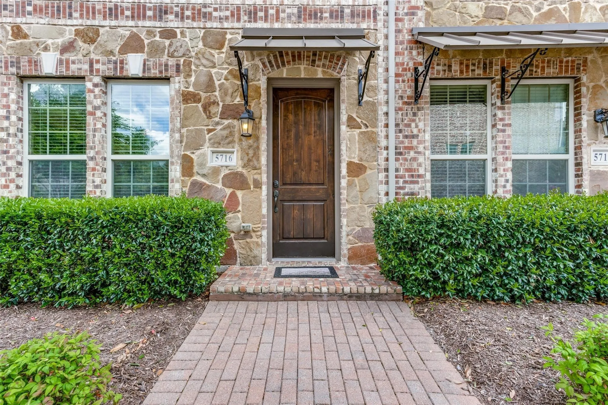 Brick and stone facade with a dark wood entry door, featuring a paved walkway, mature shrubbery, and decorative awnings