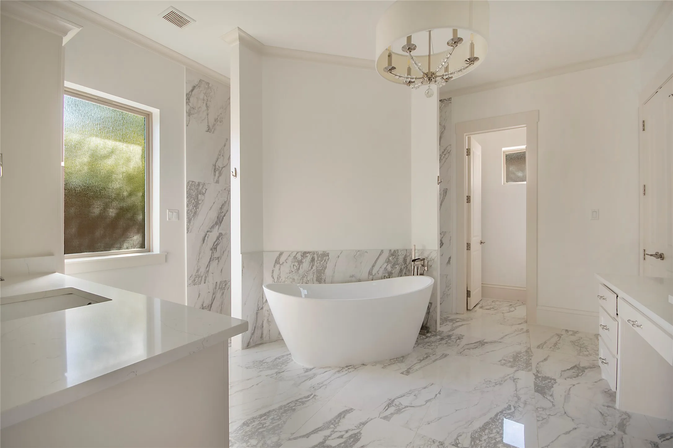 Freestanding soaking tub, white vanity with integrated sink, marble-look tile flooring, and a decorative chandelier