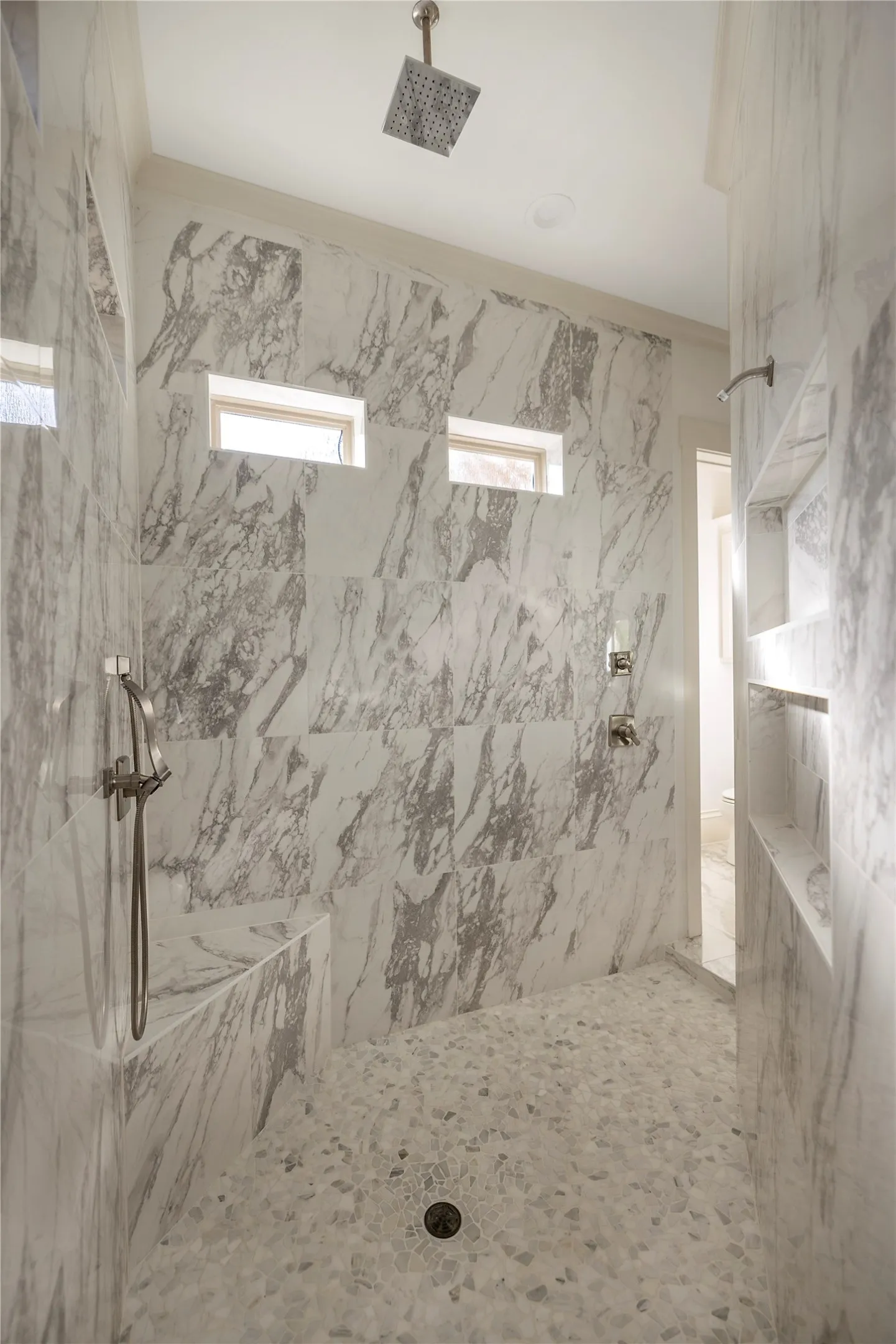 Walk-in shower featuring large format wall tiling, pebble-finish flooring, a built-in bench, a rain showerhead, and a handheld shower fixture