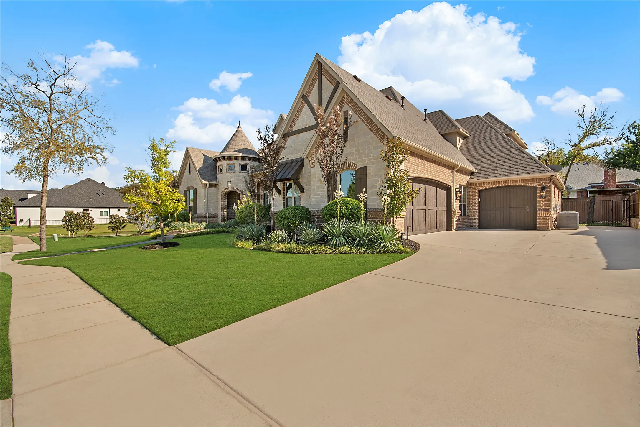 Stone and brick facade with a prominent turret, multiple gables, a well-maintained lawn, and a wide paved driveway leading to a three-car garage