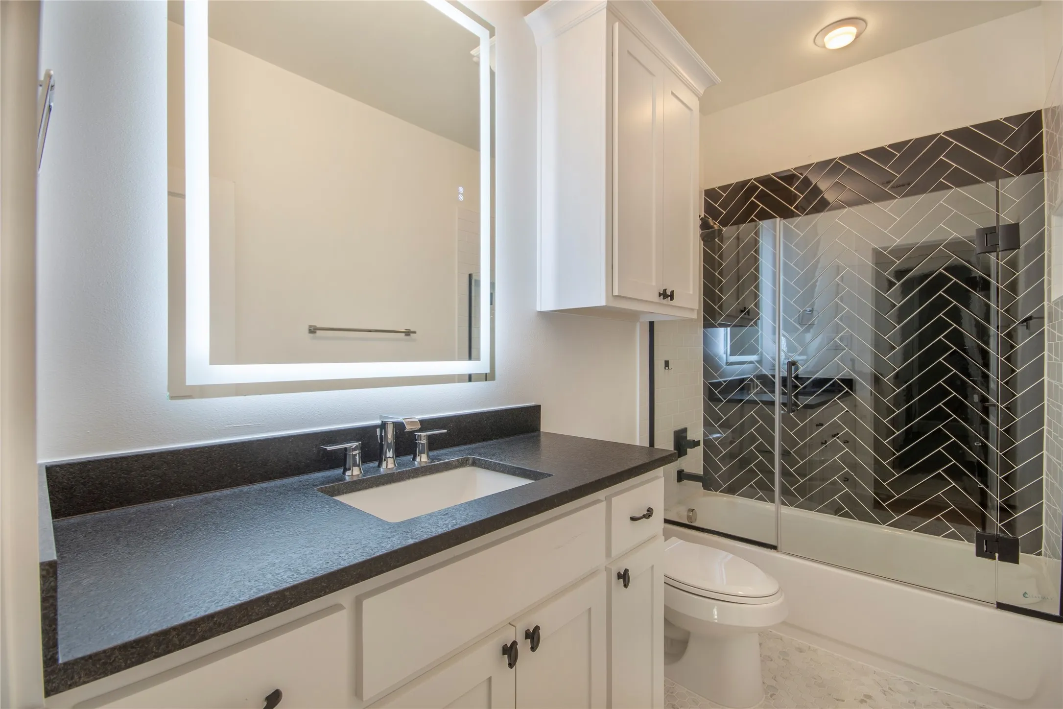 Vanity with a dark countertop and undermount sink, illuminated mirror, white cabinetry, and herringbone tile shower surround with a clear glass enclosure