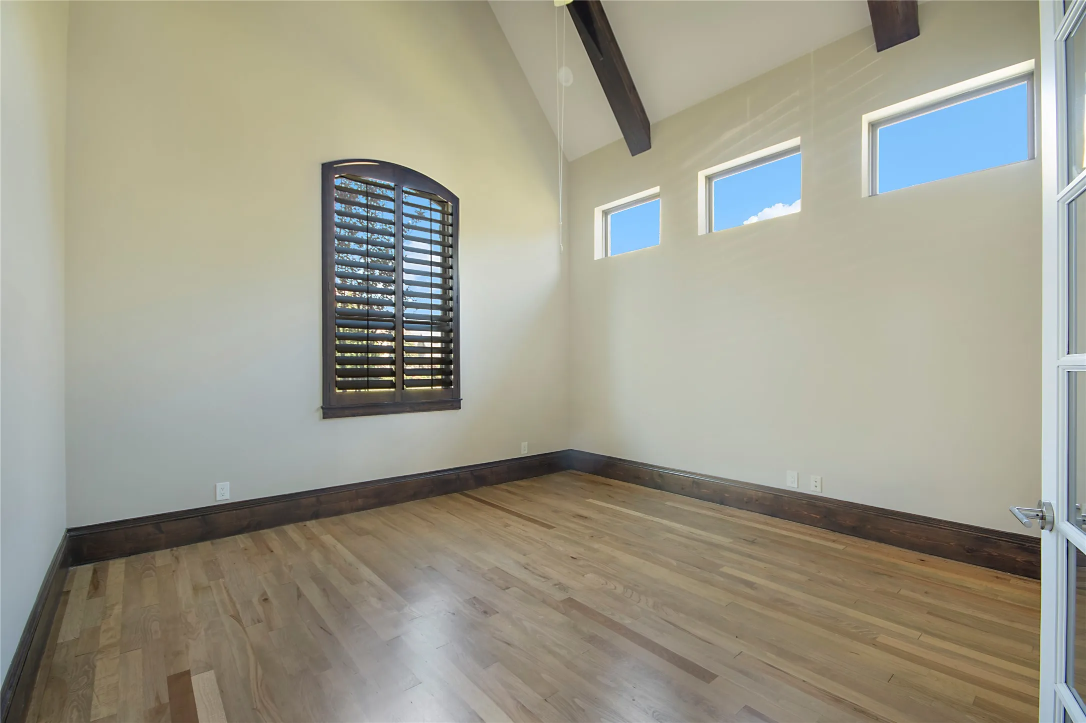 Vaulted ceiling with exposed wood beams, wood-finish flooring, dark wood baseboards, and arched window with plantation shutters