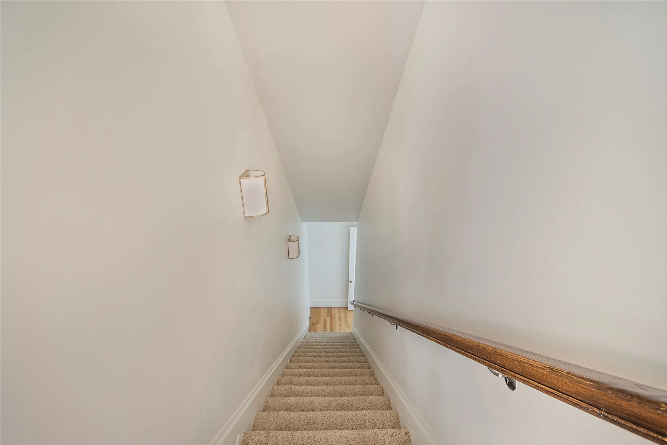 Carpeted staircase featuring a wood handrail and wall-mounted sconces