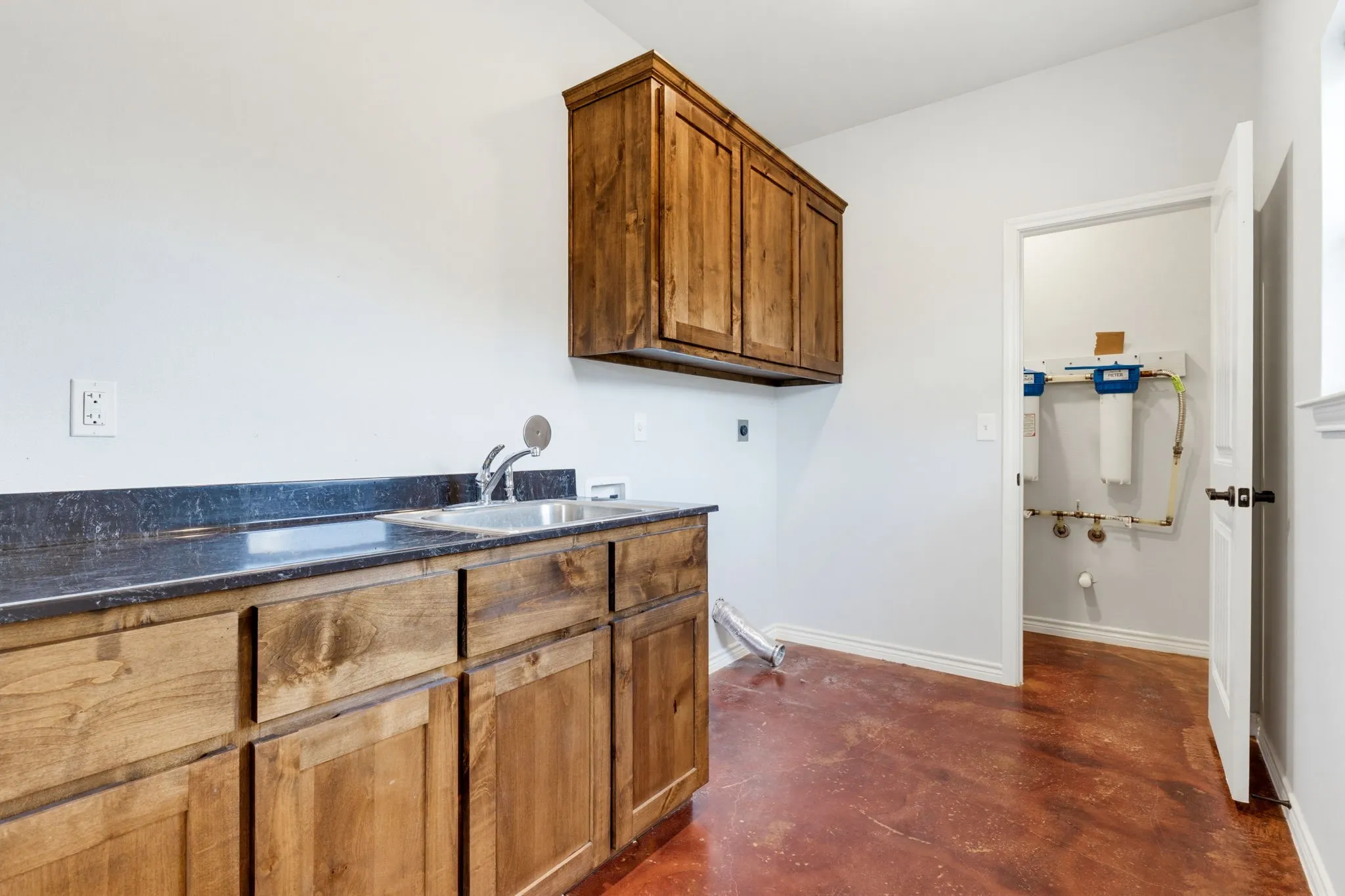 Stained concrete flooring, wood cabinetry, dark countertop, and a utility sink