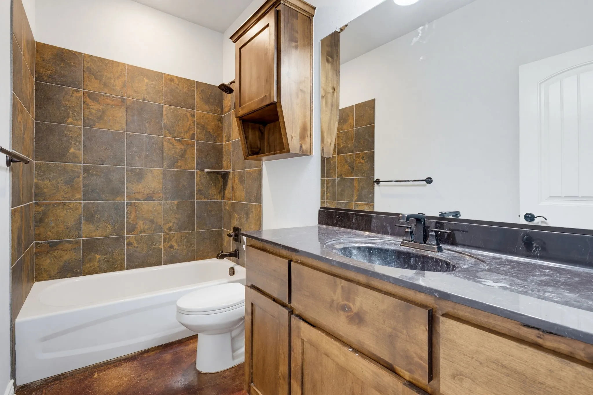 Vanity with an integrated sink and dark countertop, wood-finish cabinetry, full-width mirror, and a tub-shower combination with tiled surround