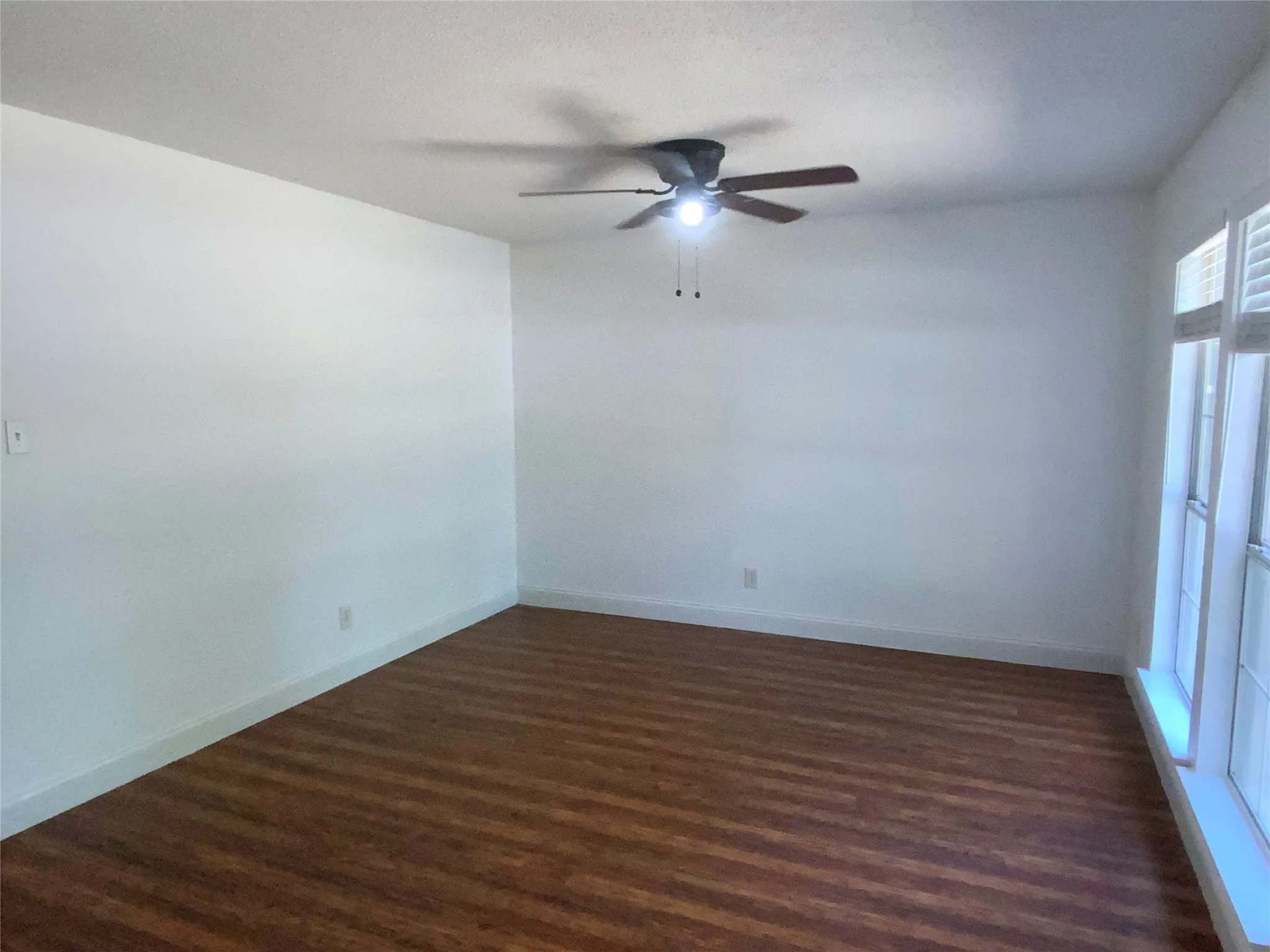 Spacious room featuring wood-finish flooring, white walls, a ceiling fan with integrated lighting, and multiple windows with white shutters