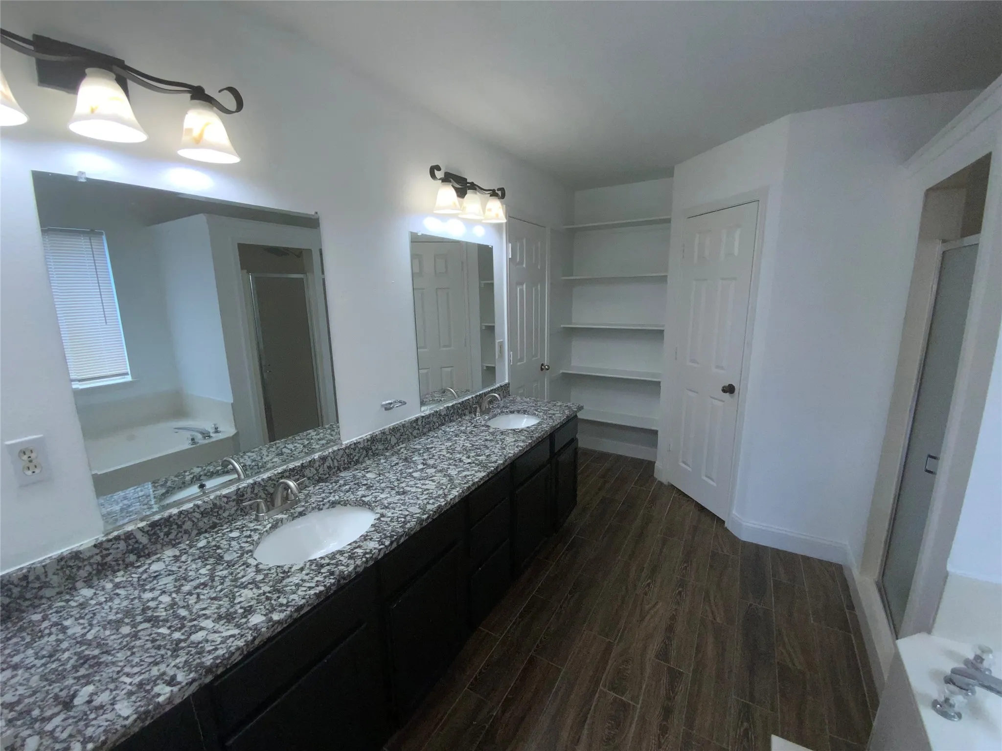 Dual vanity with granite countertop, wood-finish flooring, built-in shelving, glass-enclosed shower, and built-in tub