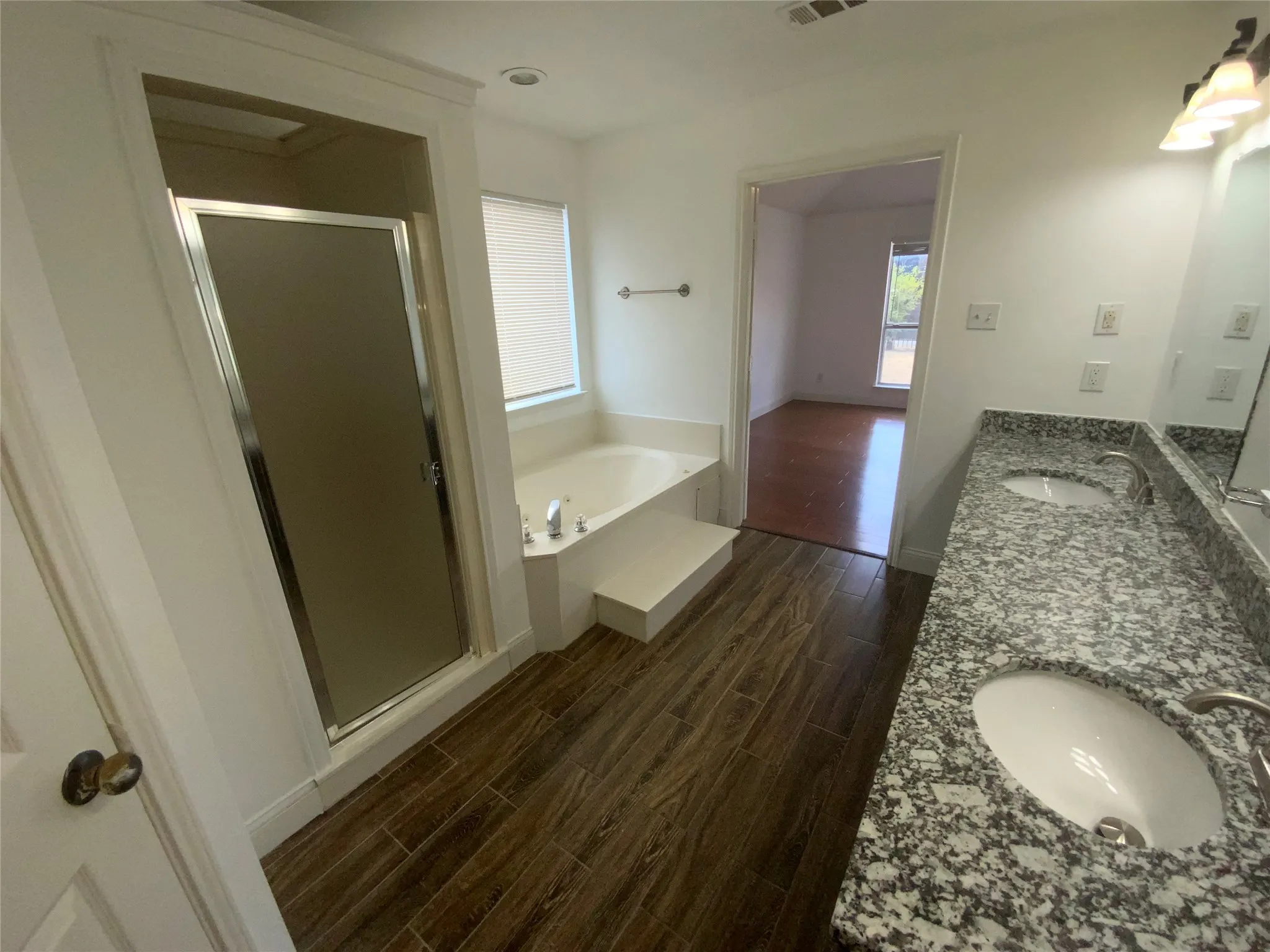 Dual vanity with undermount sinks and stone countertops, wood-look tile flooring, separate shower enclosure with frosted glass, and a built-in soaking tub