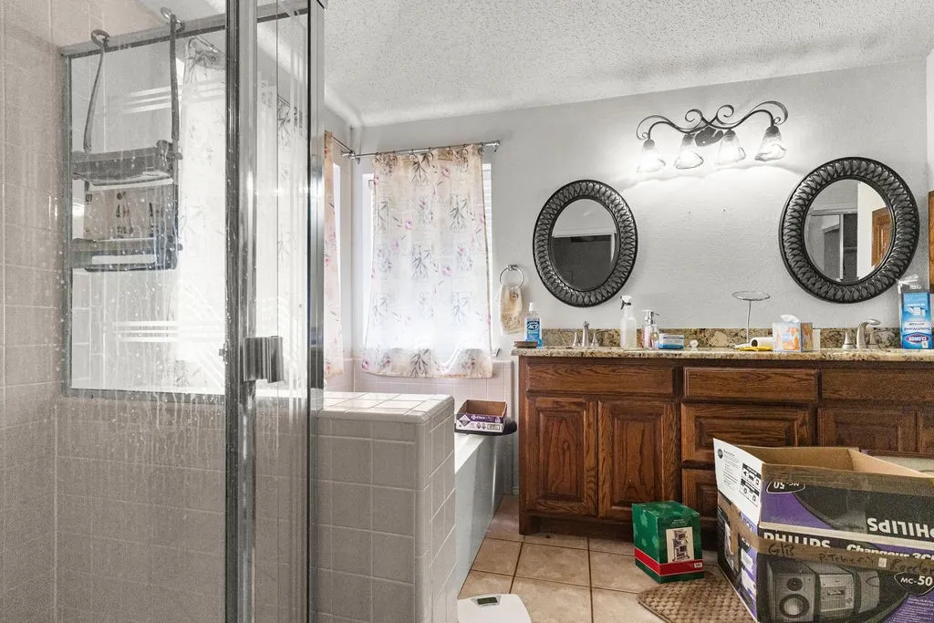Enclosed shower with glass paneling, tile-surround bathtub, double vanity with wood cabinetry, dual oval mirrors, and a four-light vanity fixture