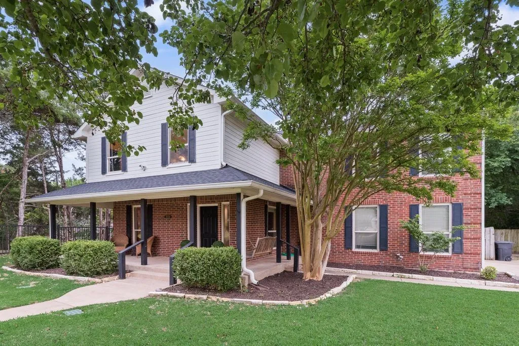 Welcoming exterior featuring a classic brick-and-siding design, mature shade trees, manicured landscaping, and a spacious covered front porch.