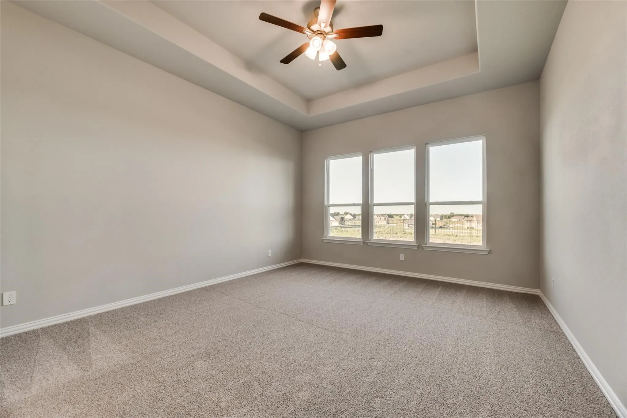 Tray ceiling with an attached ceiling fan, three large windows, and neutral carpeting