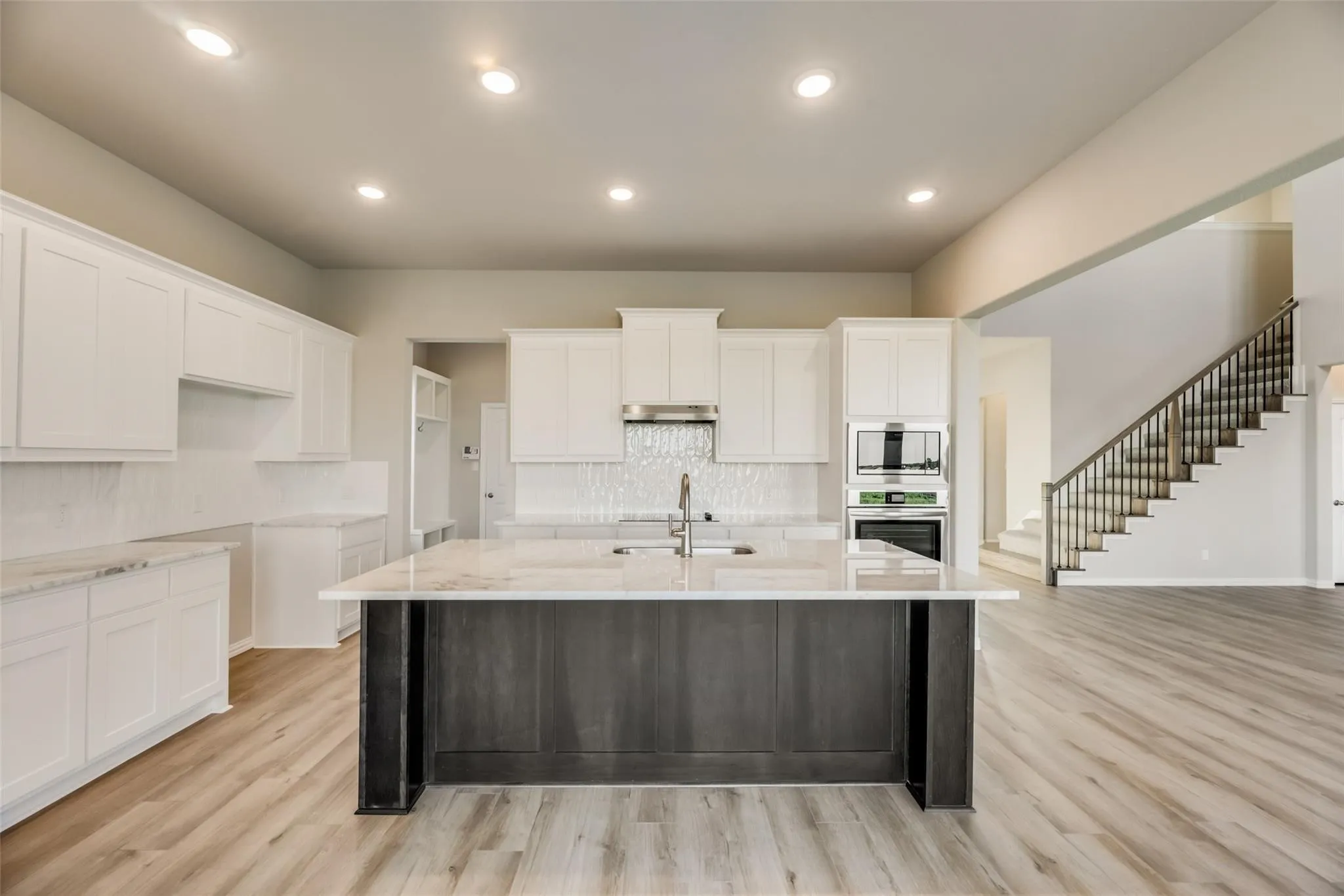 White cabinetry, stone countertops, integrated stainless steel appliances, wood-finish flooring, and a staircase with a dark wood railing