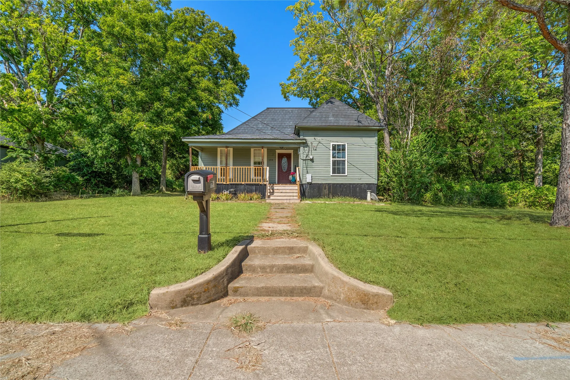 Exterior featuring light green siding, dark roof shingles, an inviting front porch with wood railings, and a concrete walkway with integrated steps