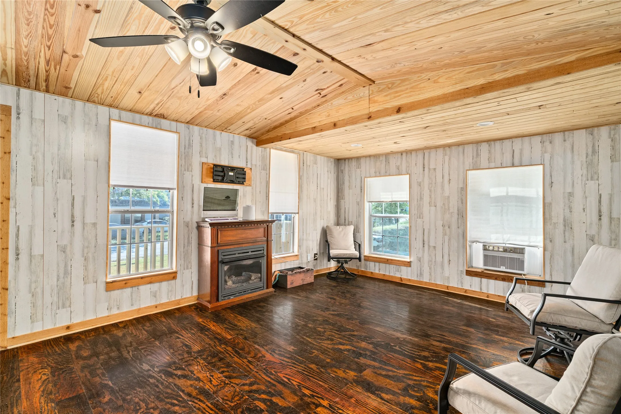 Vaulted wood-paneled ceiling, wood-finish flooring, shiplap-style wall covering, multiple windows with roller shades, and a built-in fireplace