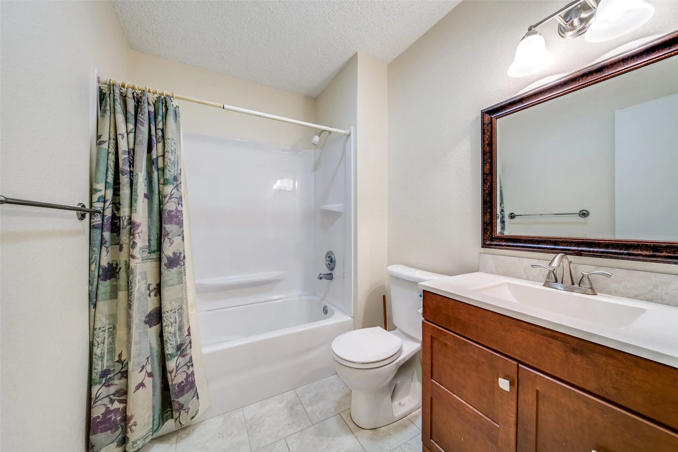 White shower/tub combination, wood-finish vanity with built-in sink, framed mirror, and tile flooring