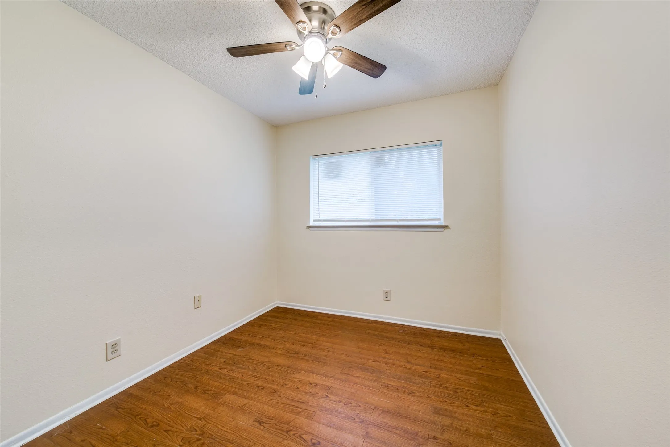 Wood-finish flooring, textured walls, window with a roller shade, and ceiling fan with integrated lighting
