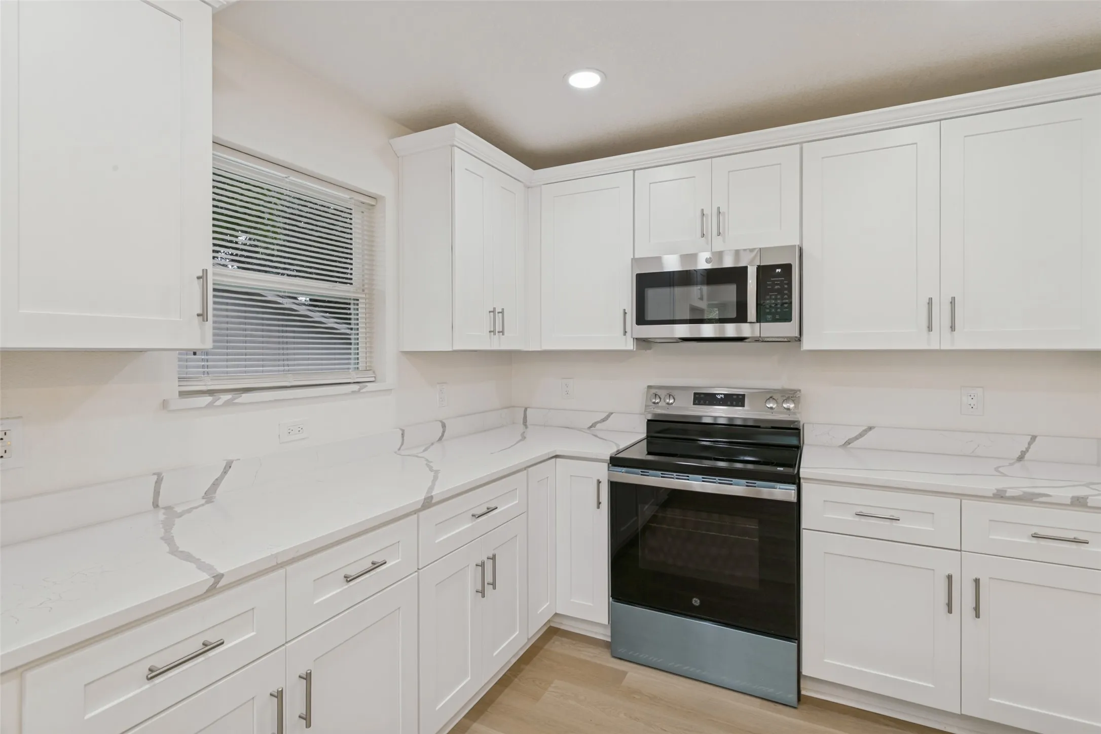 White shaker-style cabinetry, stainless steel microwave, stainless steel range, light-toned wood-finish flooring, and light-toned countertops with veining