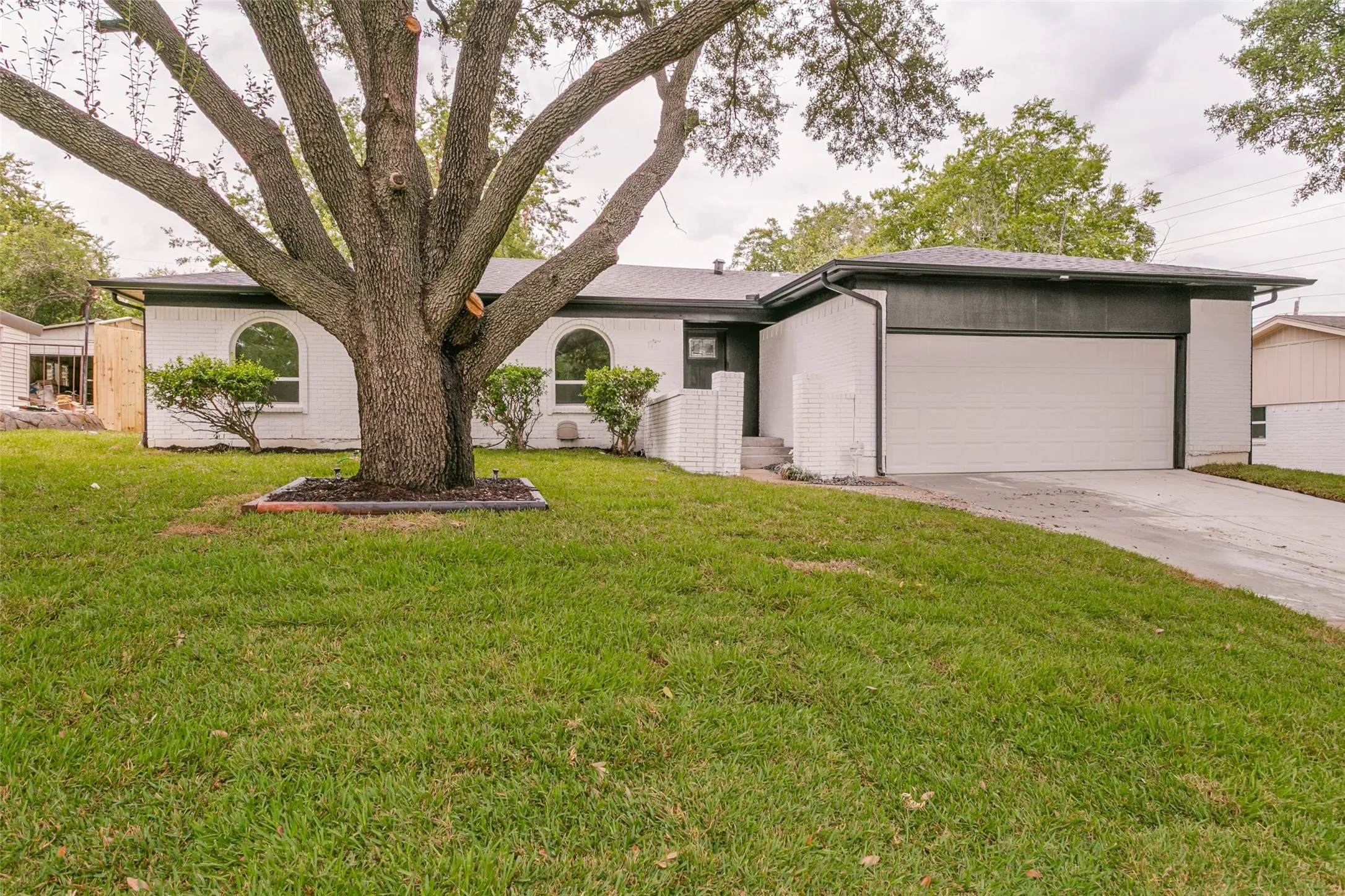 White brick exterior with dark trim, prominent mature tree, arched windows, and an attached two-car garage