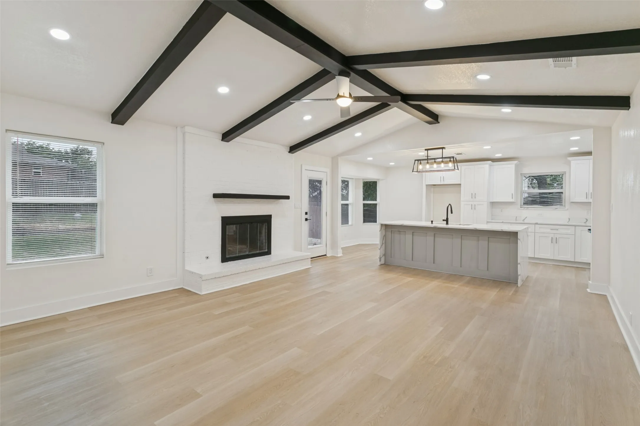 Vaulted ceiling with exposed beams, recessed lighting, wood-finish flooring, white brick fireplace, and kitchen island with countertop