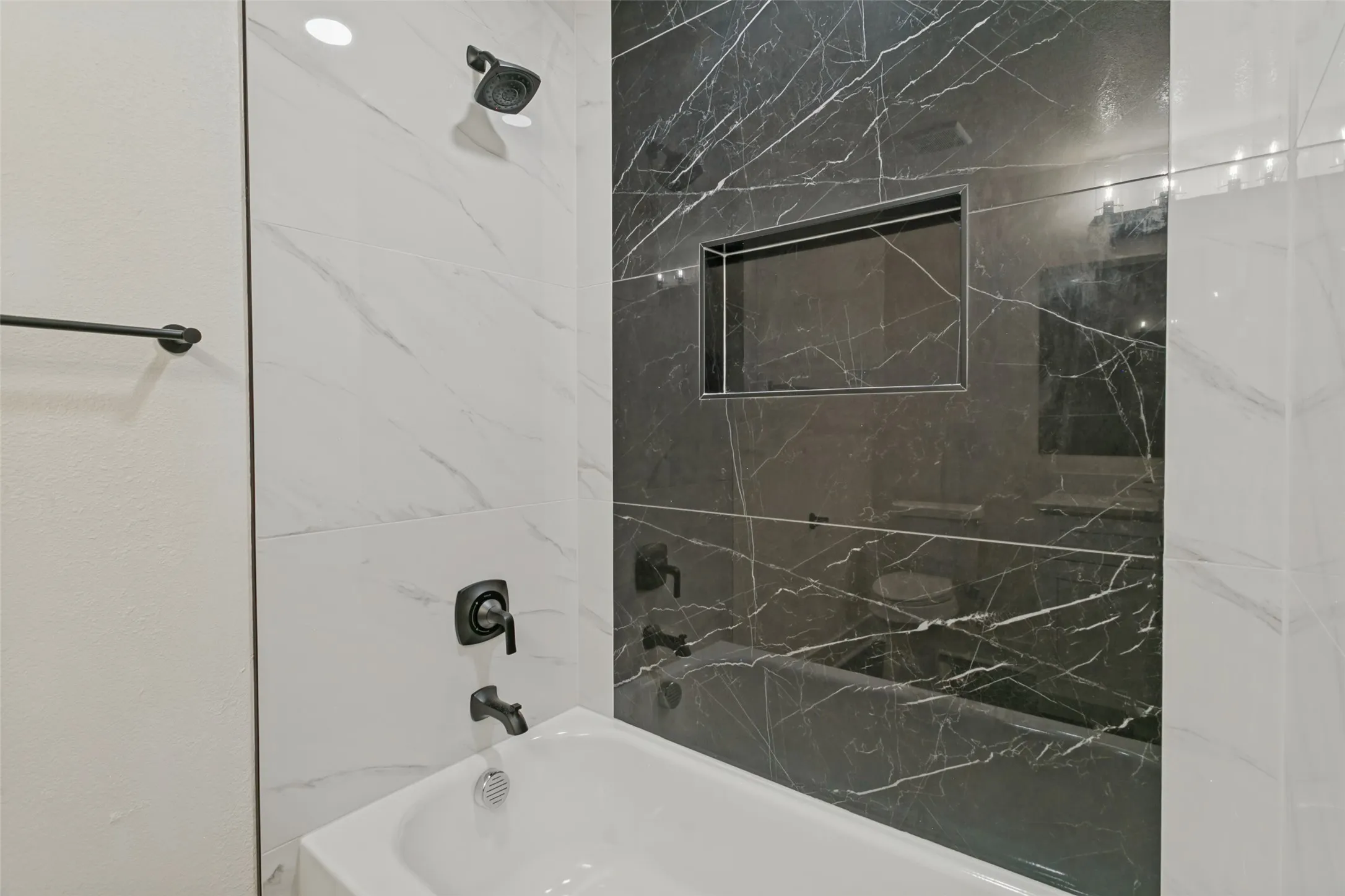 White bathtub with matte black fixtures, dark large-format tile with light veining, white large-format tile with light veining, and a recessed shower niche
