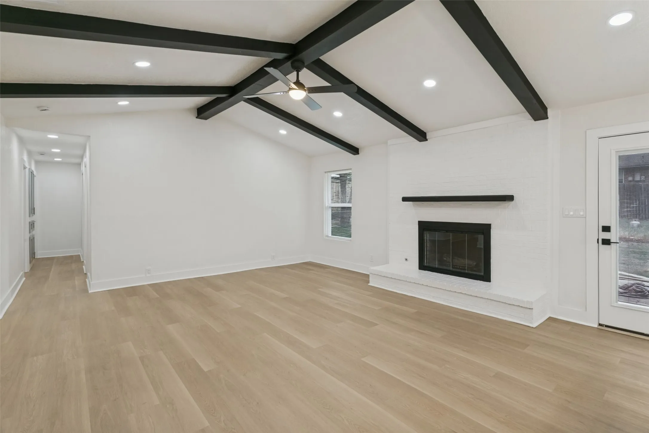 Vaulted ceiling with exposed black beams, light wood-finish flooring, white brick fireplace with black mantel, recessed lighting, and a glass-paned exterior door