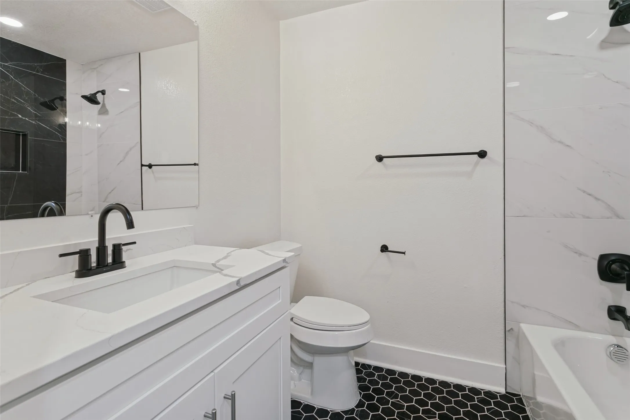 Modern bathroom featuring a white shaker-style vanity with integrated sink and quartz countertop, matte black faucet and hardware, frameless glass shower enclosure with subway tile surround, hexagonal tile flooring in black and white, and a toilet with chrome fixtures