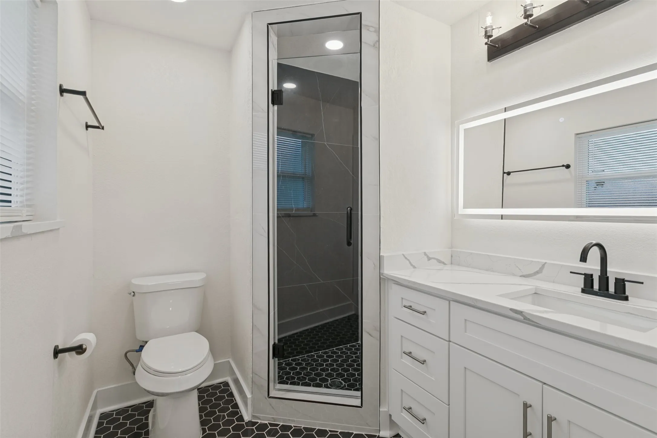 Frameless glass shower enclosure, dual-sink vanity with white cabinetry, white stone-finish countertop, black hexagonal floor tiling, and an illuminated mirror