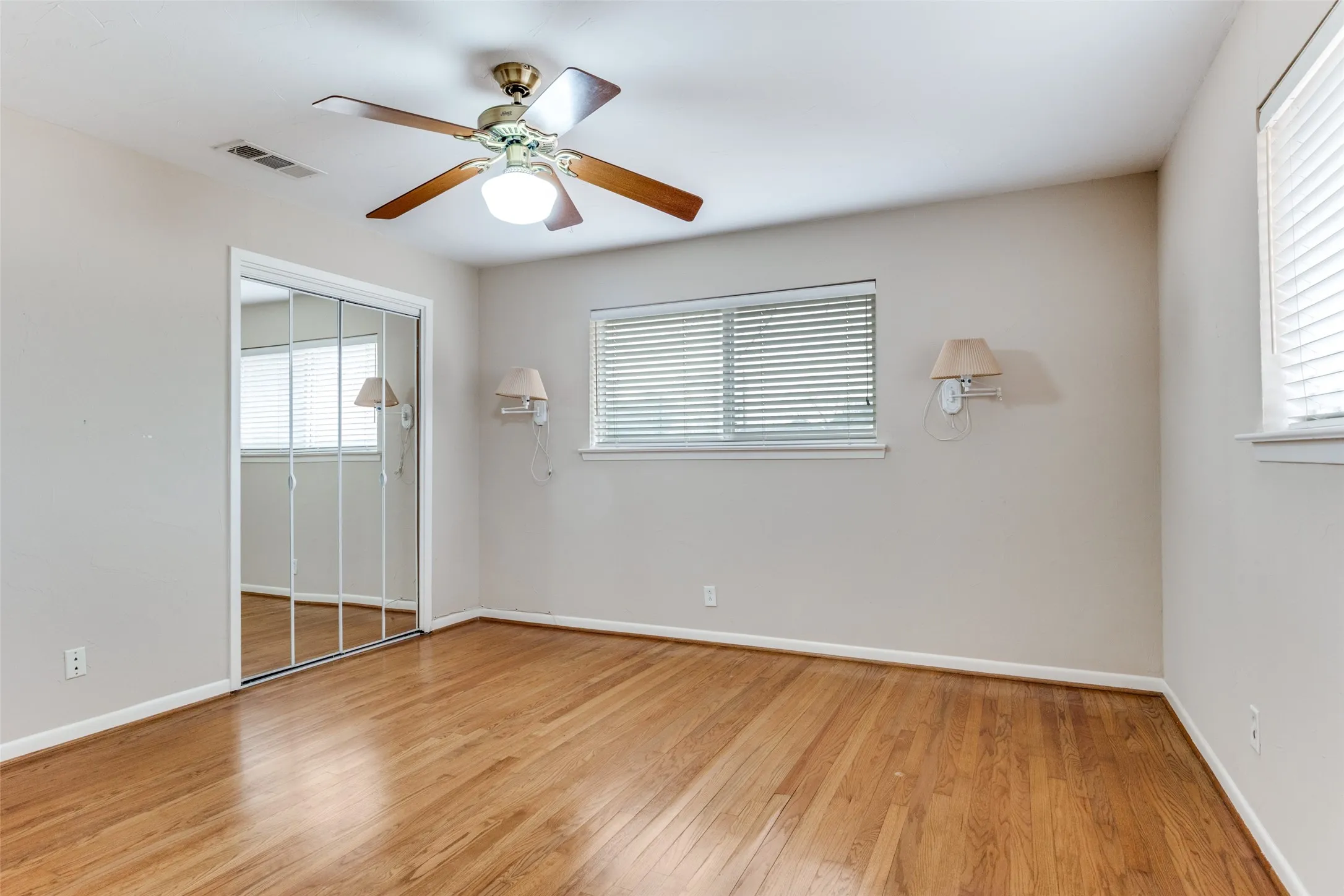 Hardwood flooring, mirrored closet doors, ceiling fan with light fixture, window treatments, and wall-mounted sconces