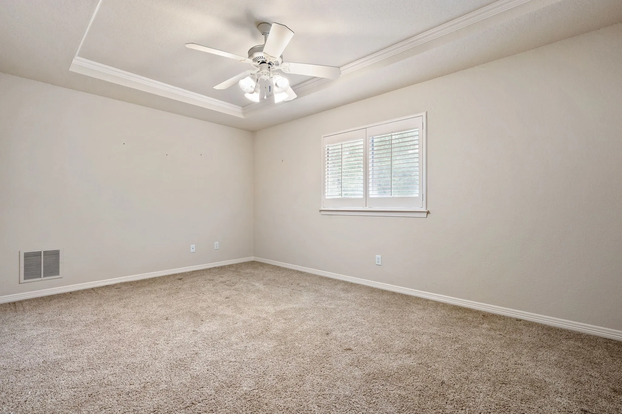 Tray ceiling with crown molding, ceiling fan with integrated lighting, window with white plantation shutters, and light-colored carpet flooring