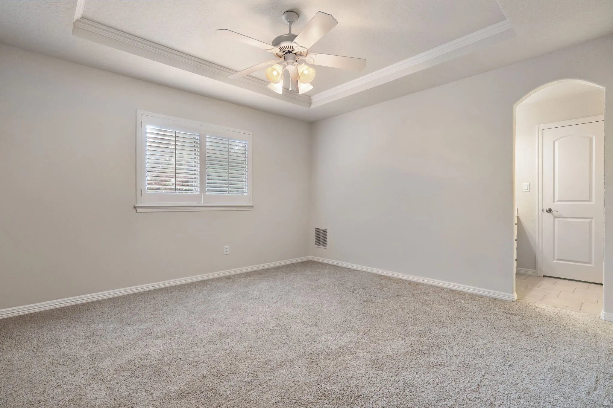 Tray ceiling with a ceiling fan, window with plantation shutters, light-toned carpet flooring, and an arched doorway