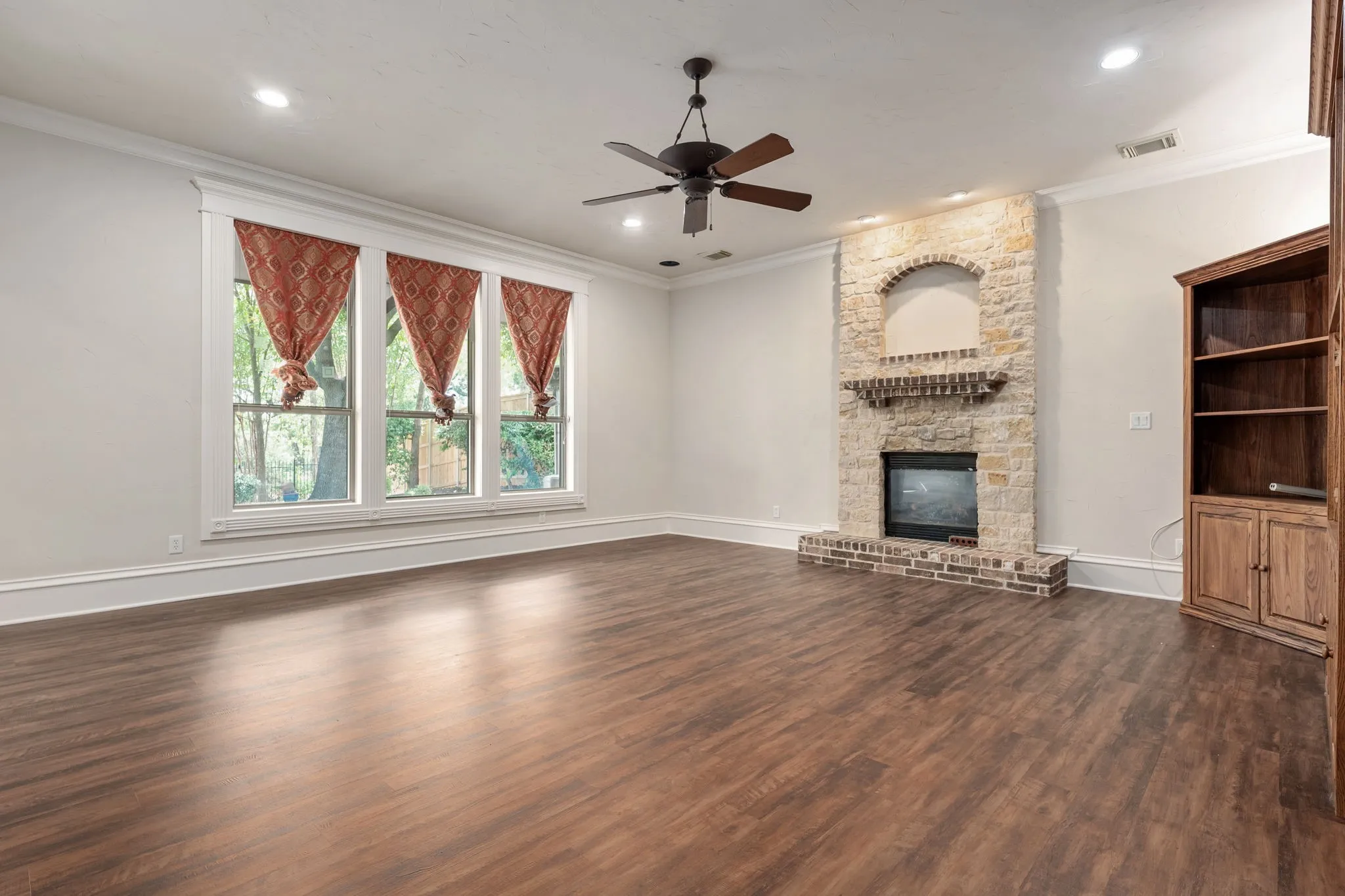 Wood-finish flooring, stone-clad fireplace with brick hearth, built-in shelving, multi-panel windows, and a ceiling fan with integrated lighting