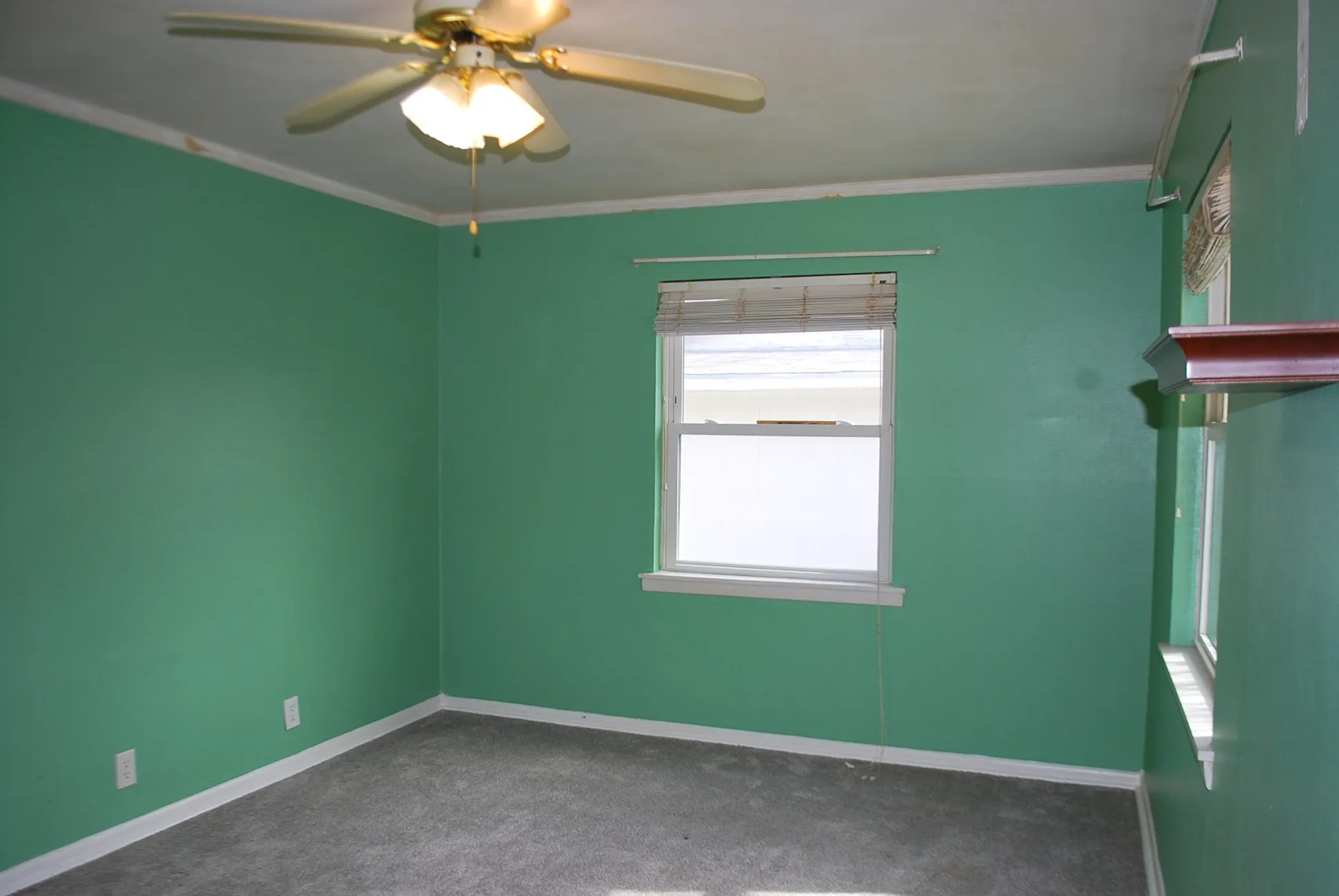 Carpeted room featuring a ceiling fan with light fixture, window with blinds, and a decorative wall shelf