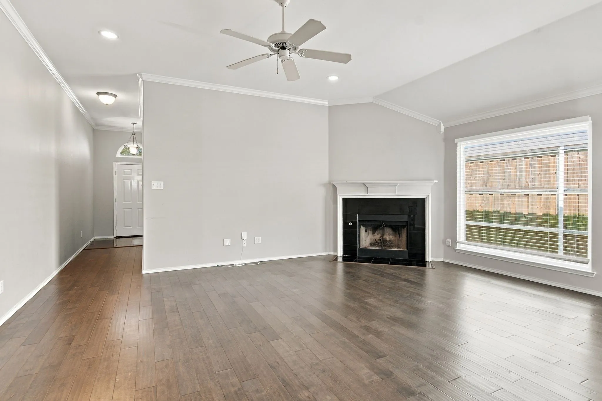 Spacious living area featuring wood-finish flooring, a fireplace with a dark surround and white mantel, a large window with blinds, and a ceiling fan