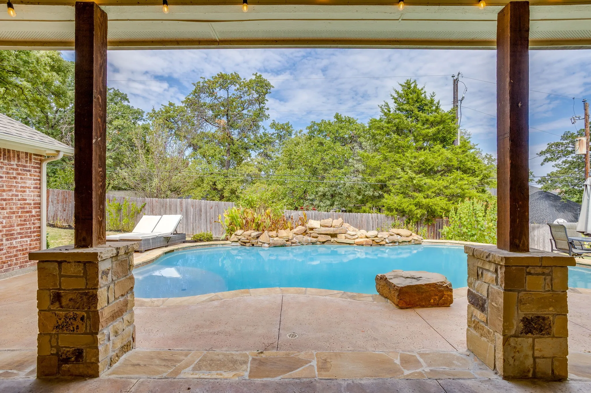Covered patio featuring stone-clad columns and wood beams, overlooking a freeform swimming pool with a rock waterfall feature and surrounding concrete decking