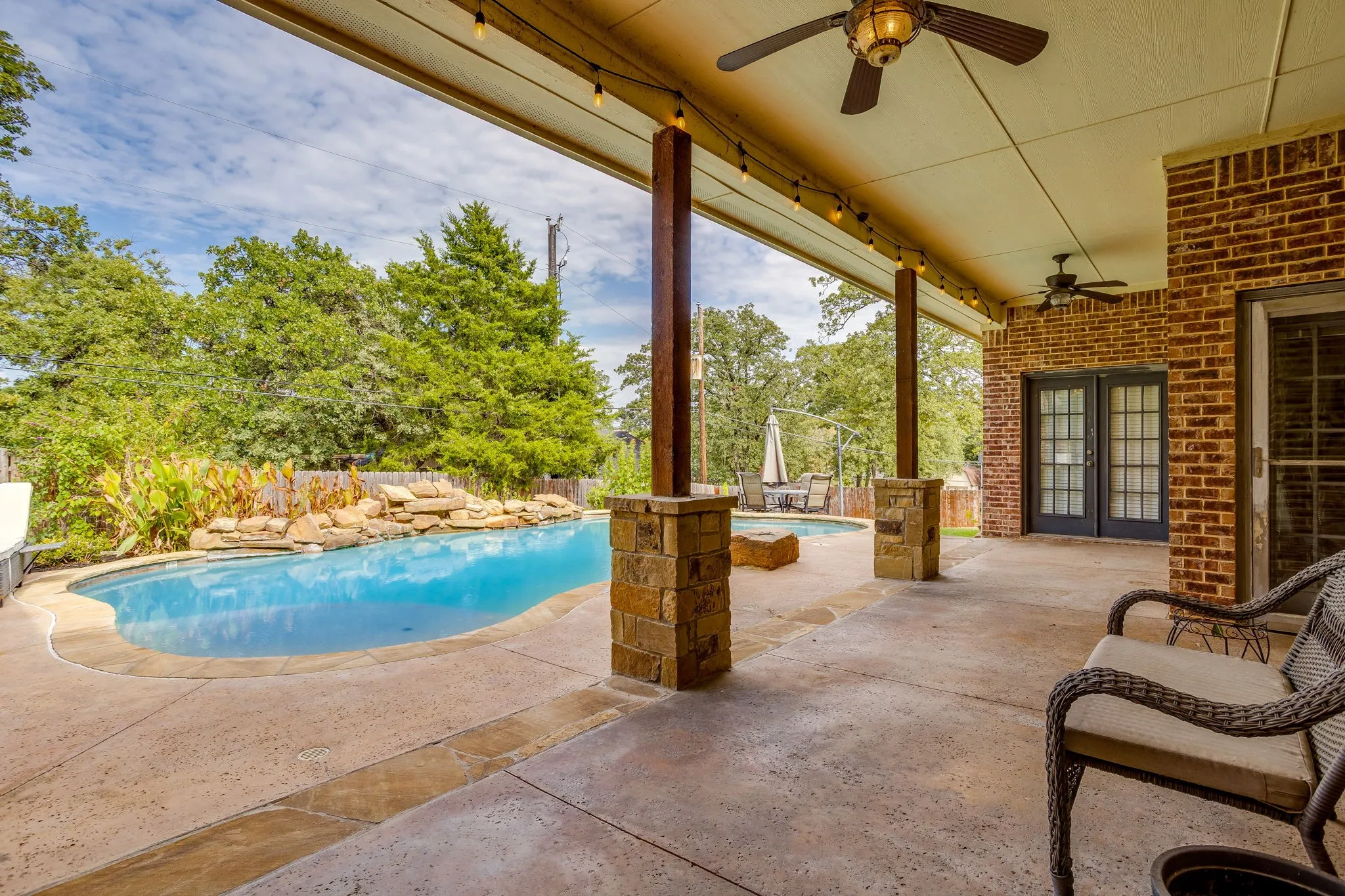 Covered patio with multiple ceiling fans, exposed brick exterior, and a built-in swimming pool with a rock waterfall feature