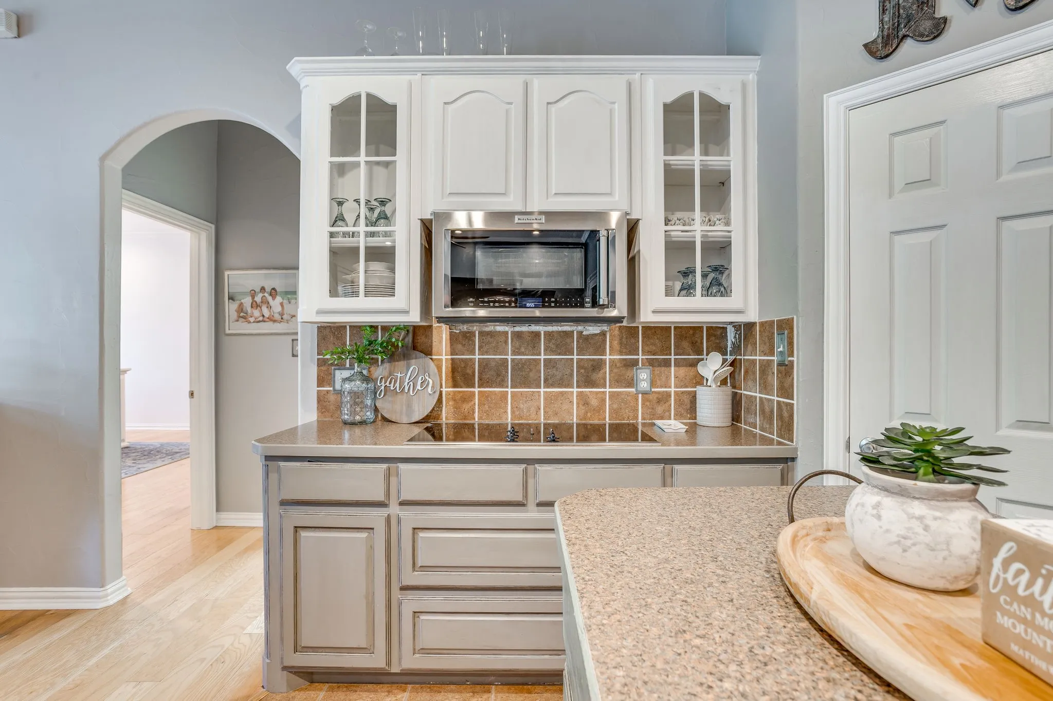 Built-in stainless steel microwave, white upper cabinetry with glass-front doors, tiled backsplash, light-colored lower cabinetry, and wood-finish flooring