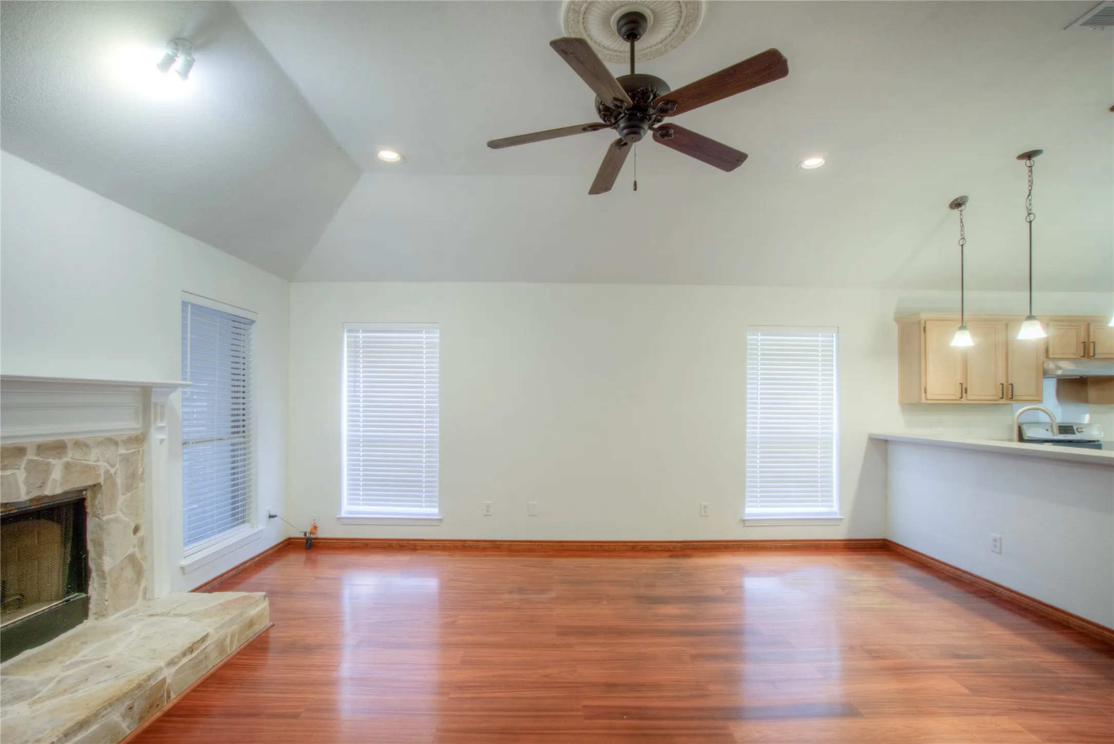 Vaulted ceiling with a decorative medallion and ceiling fan, stone-clad fireplace, wood-finish flooring, recessed lighting, and kitchen with pendant lighting