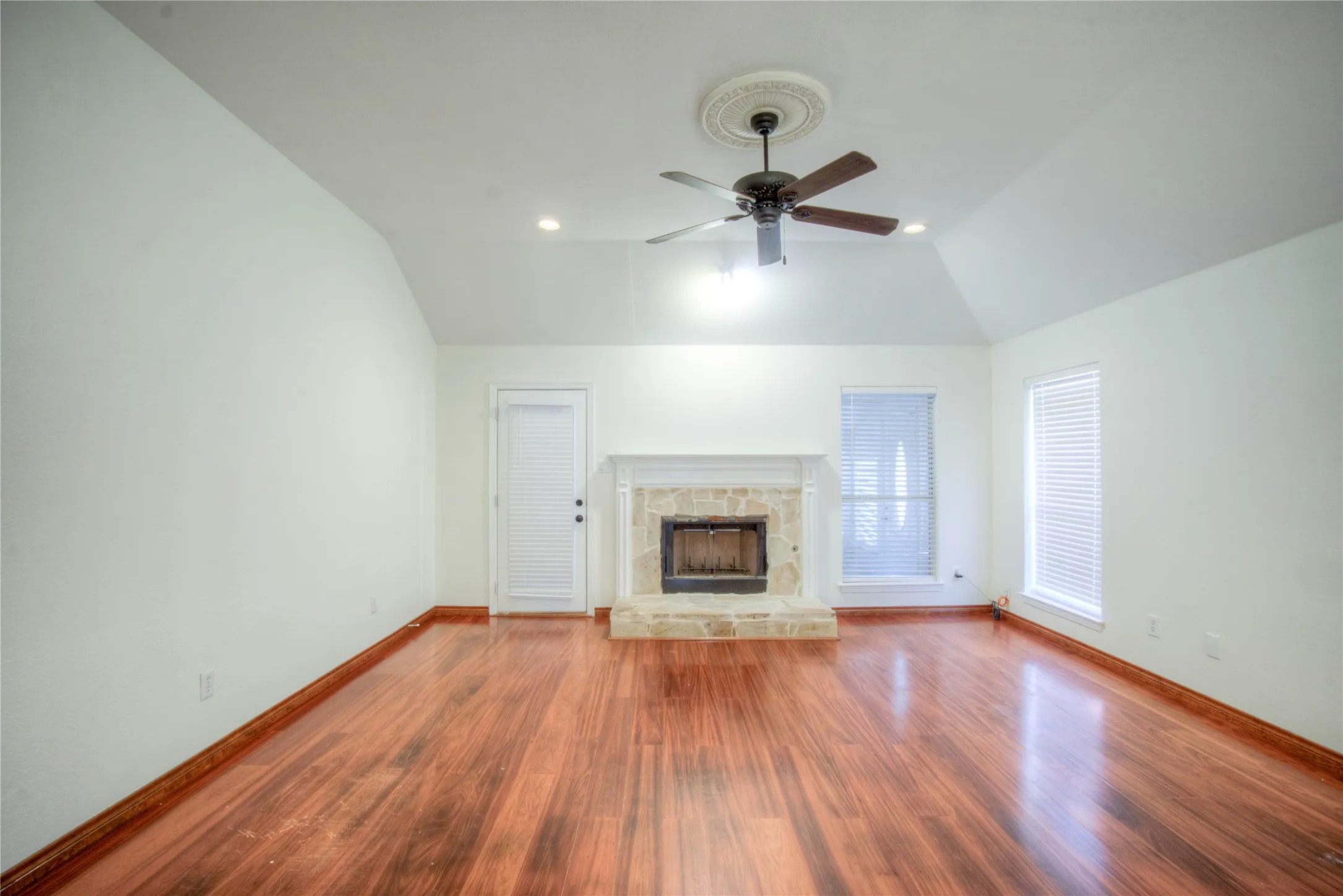 Vaulted ceiling with a decorative ceiling medallion, wood-finish flooring, stone-faced fireplace with a white mantel, and a ceiling fan
