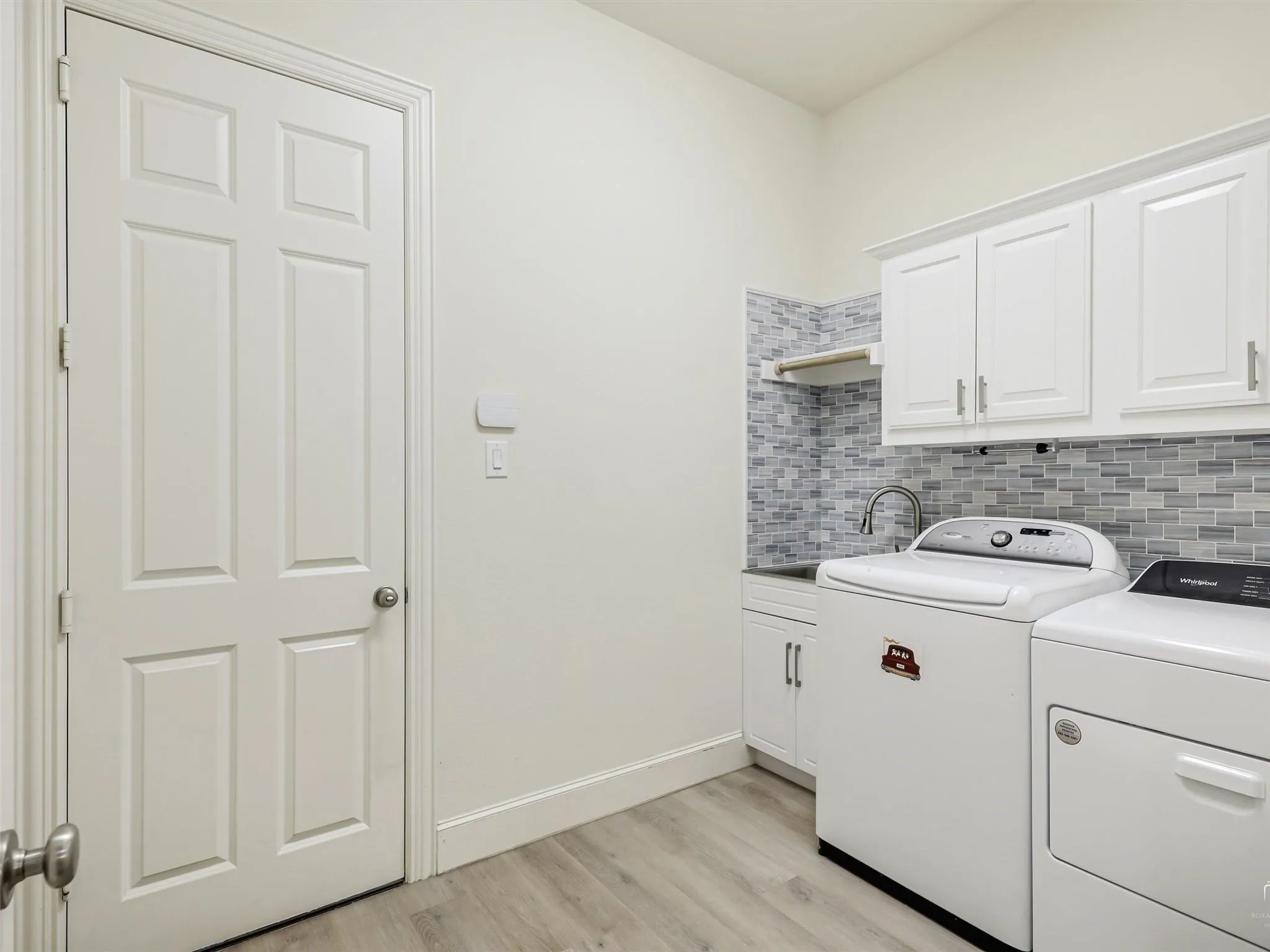Wood-finish flooring, white paneled door, white shaker-style cabinetry, mosaic tile backsplash, and a utility sink