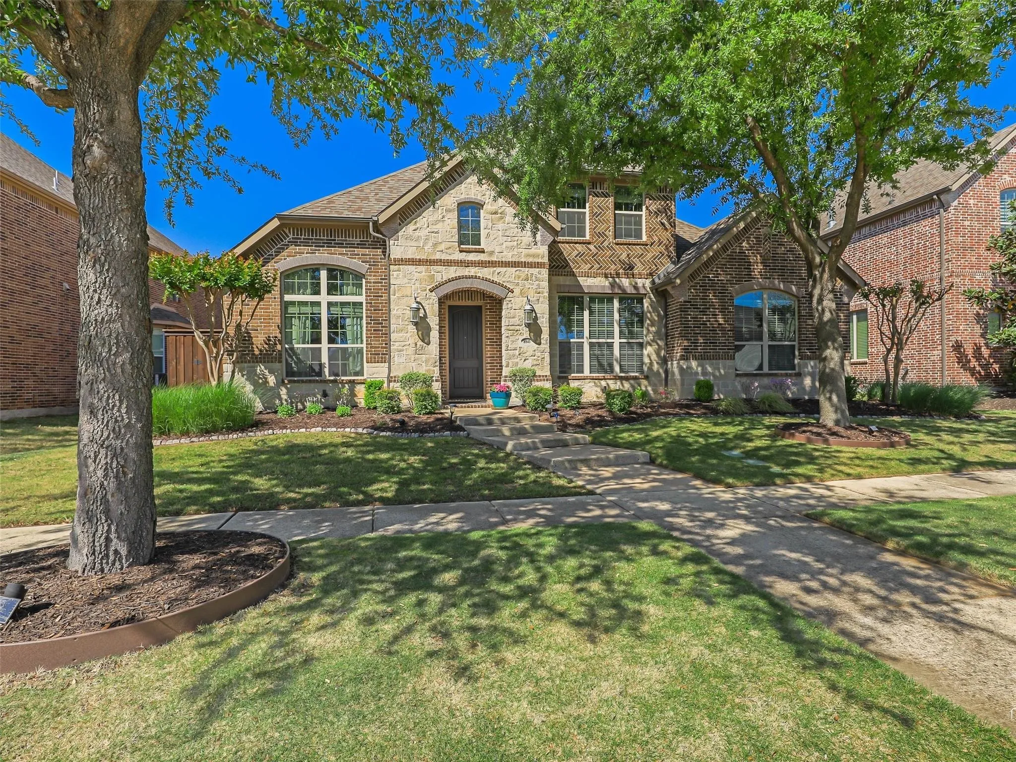 Brick and stone exterior featuring an arched entryway, manicured landscaping, and mature trees