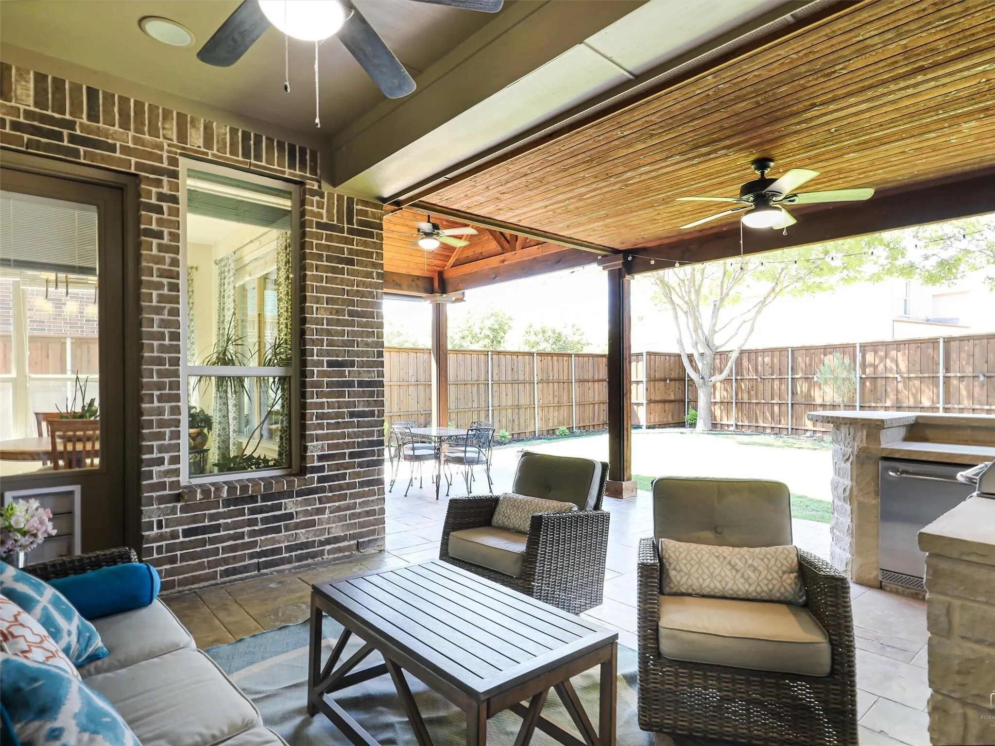 Spacious outdoor living area with a stone and brick built-in grill station, ceiling fans, a wood-slat ceiling, and a fully fenced yard