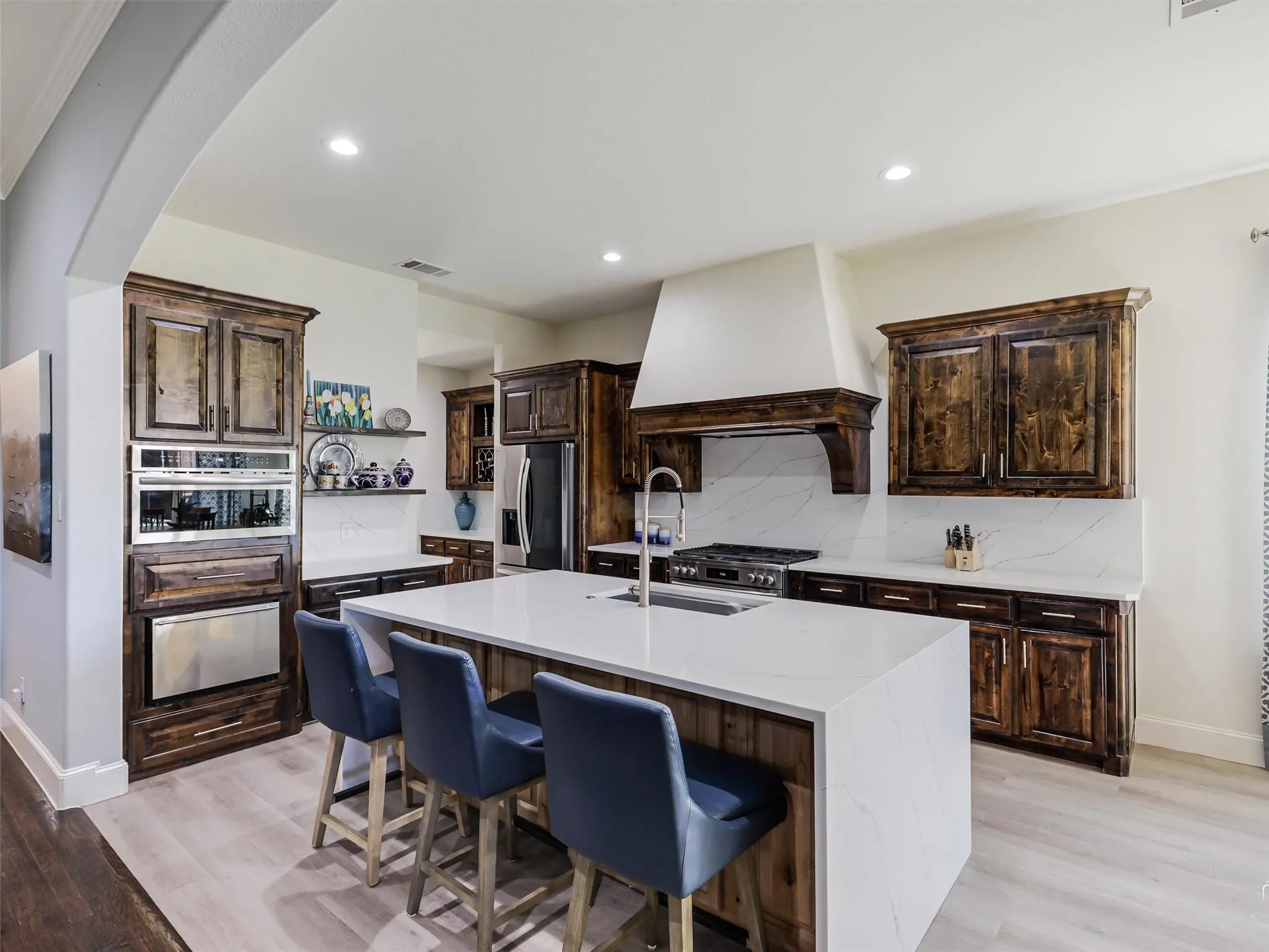 Spacious kitchen featuring an expansive island with seating, built-in stainless steel appliances, dark wood cabinetry, and light-toned countertops with a matching backsplash