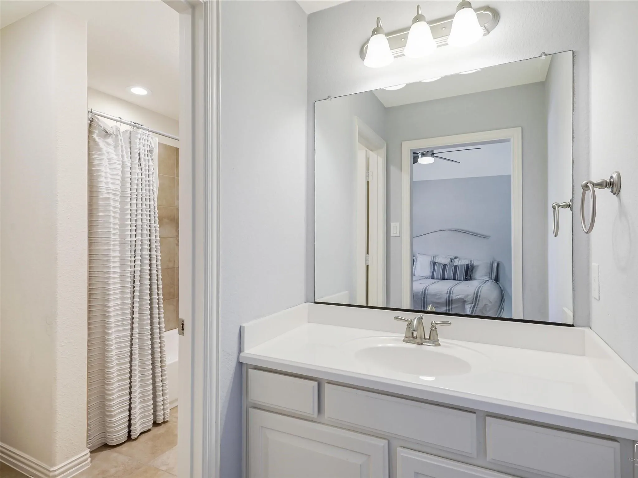 White vanity with an integrated sink, brushed nickel faucet, large wall mirror, three-light vanity fixture, and tiled shower enclosure