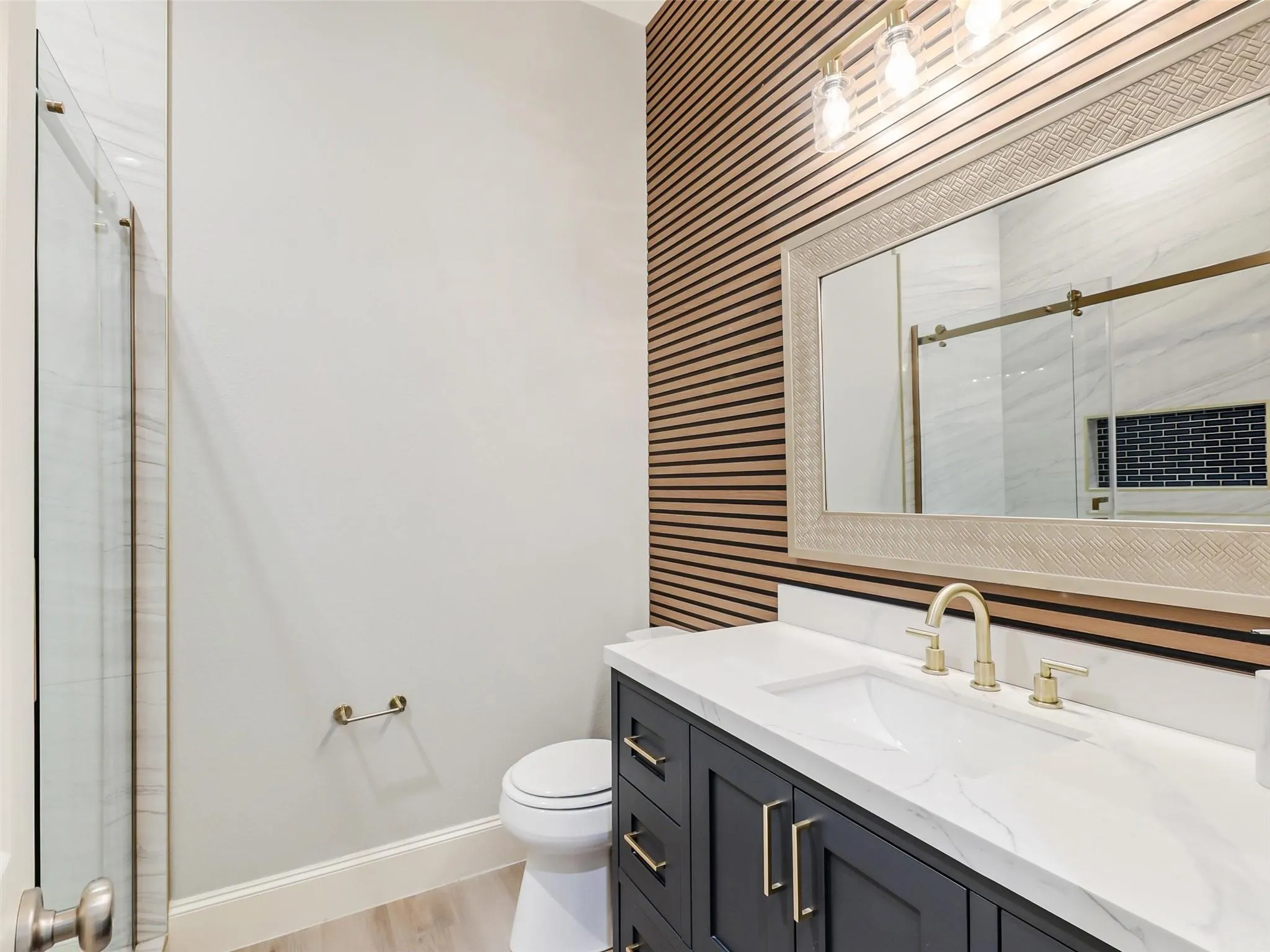 Vanity with a white countertop and gold-toned fixtures, wood-slat accent wall, framed mirror, and a glass-enclosed shower