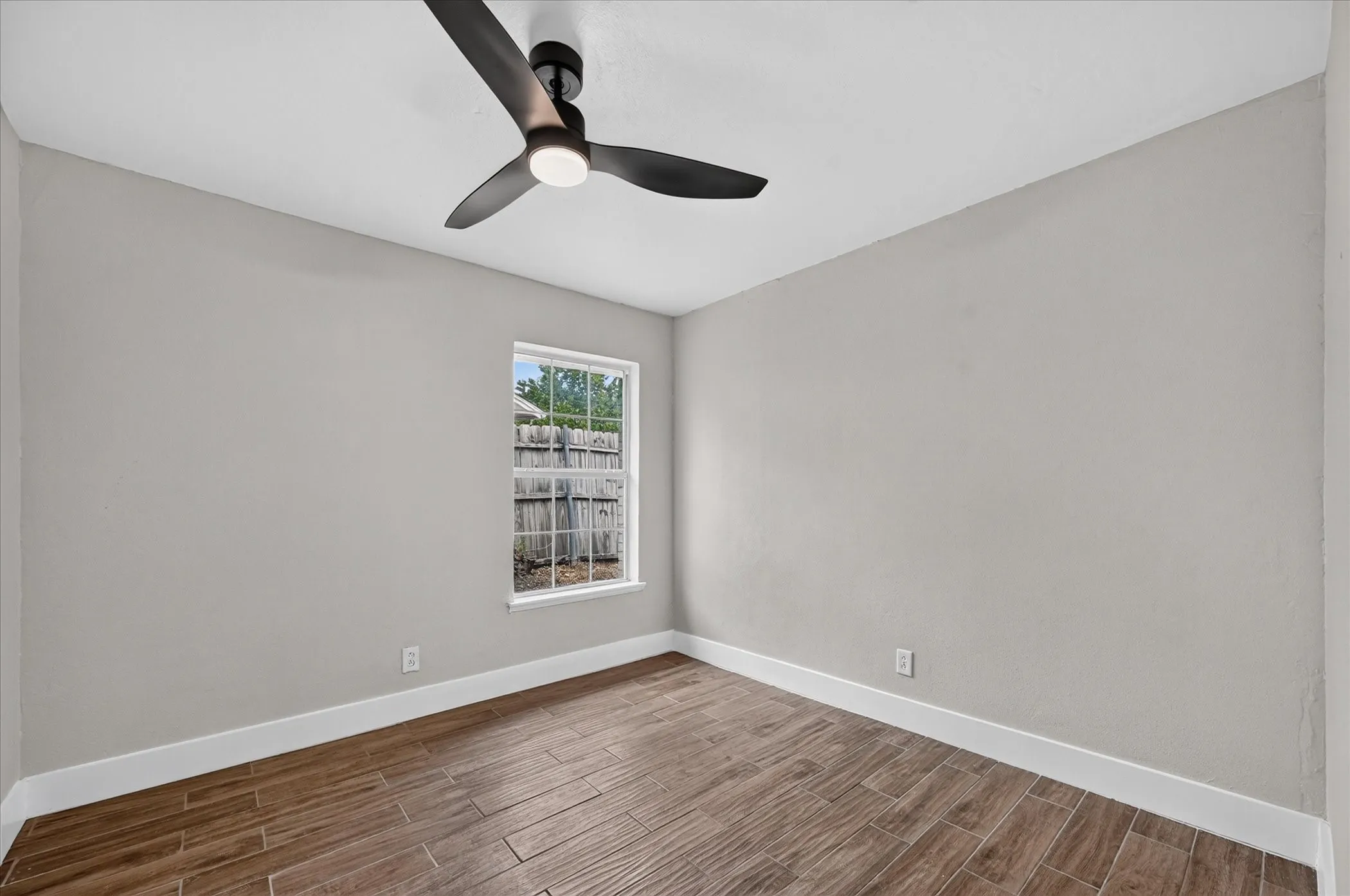 Wood-finish flooring, single window, white trim, light gray walls, and a modern black ceiling fan